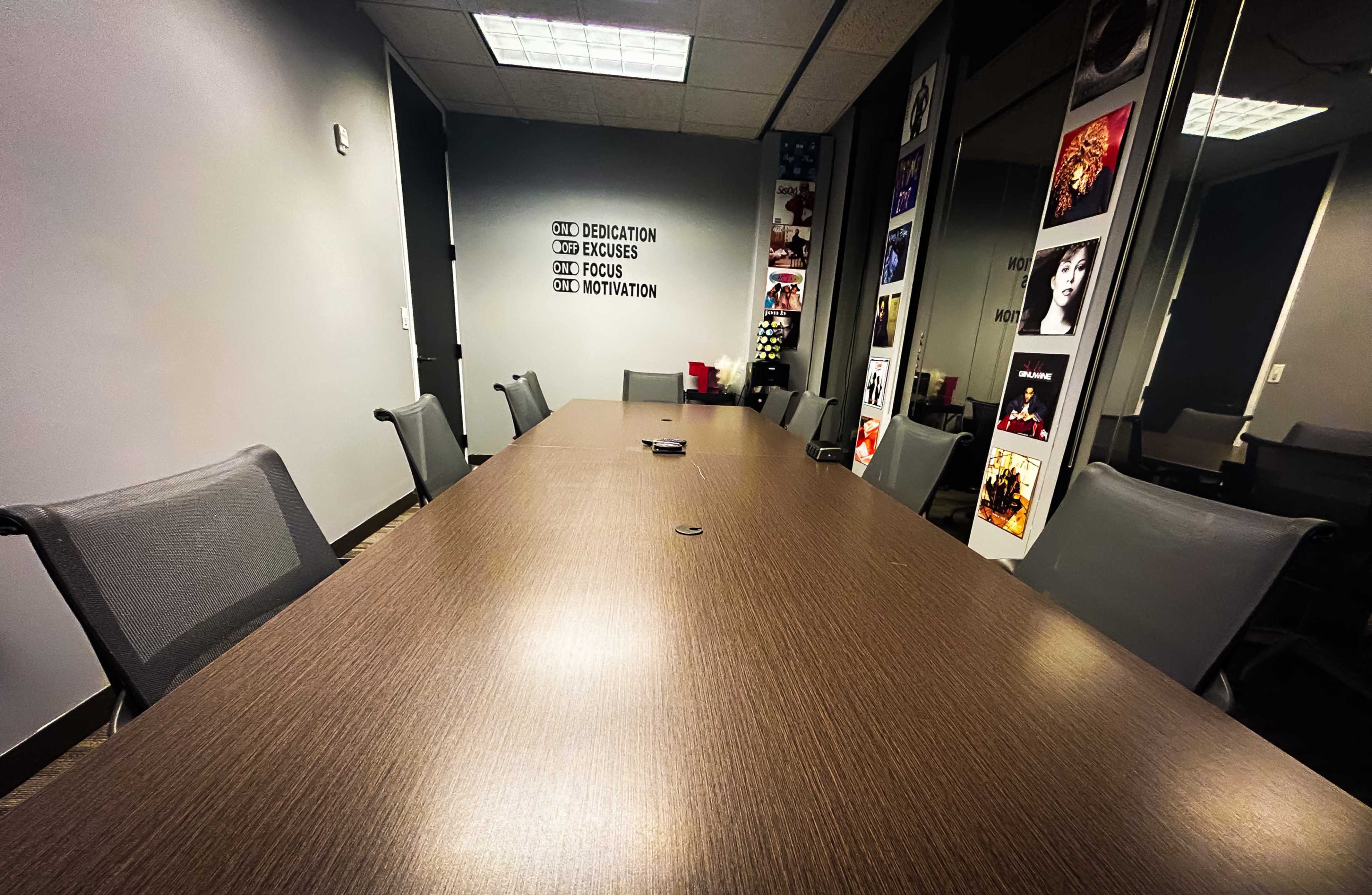A long wooden conference table surrounded by gray chairs is set in a meeting room with walls displaying framed posters.
