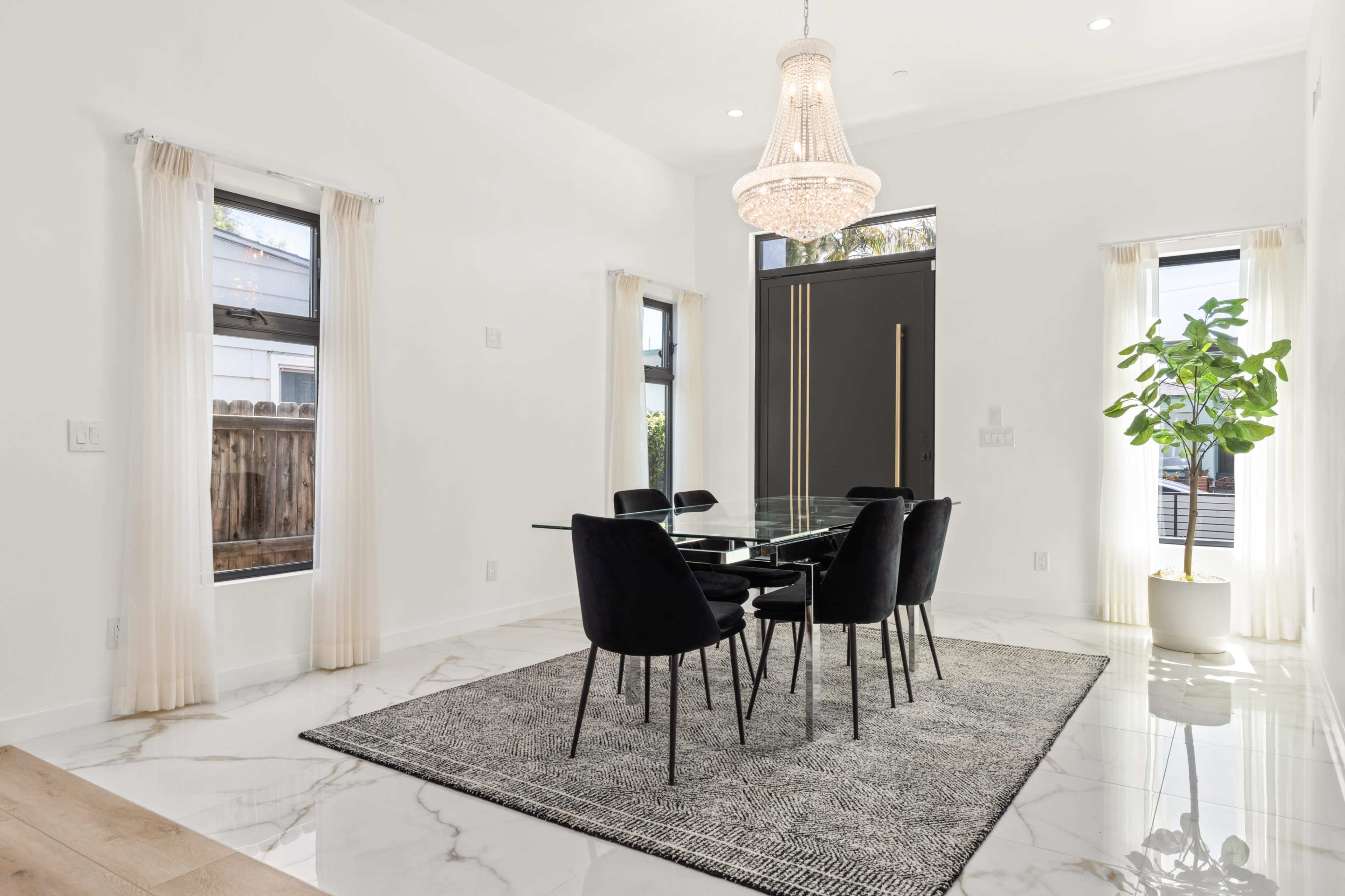 A modern dining area features a glass table surrounded by black chairs, brightened by natural light through large windows and a chandelier above.