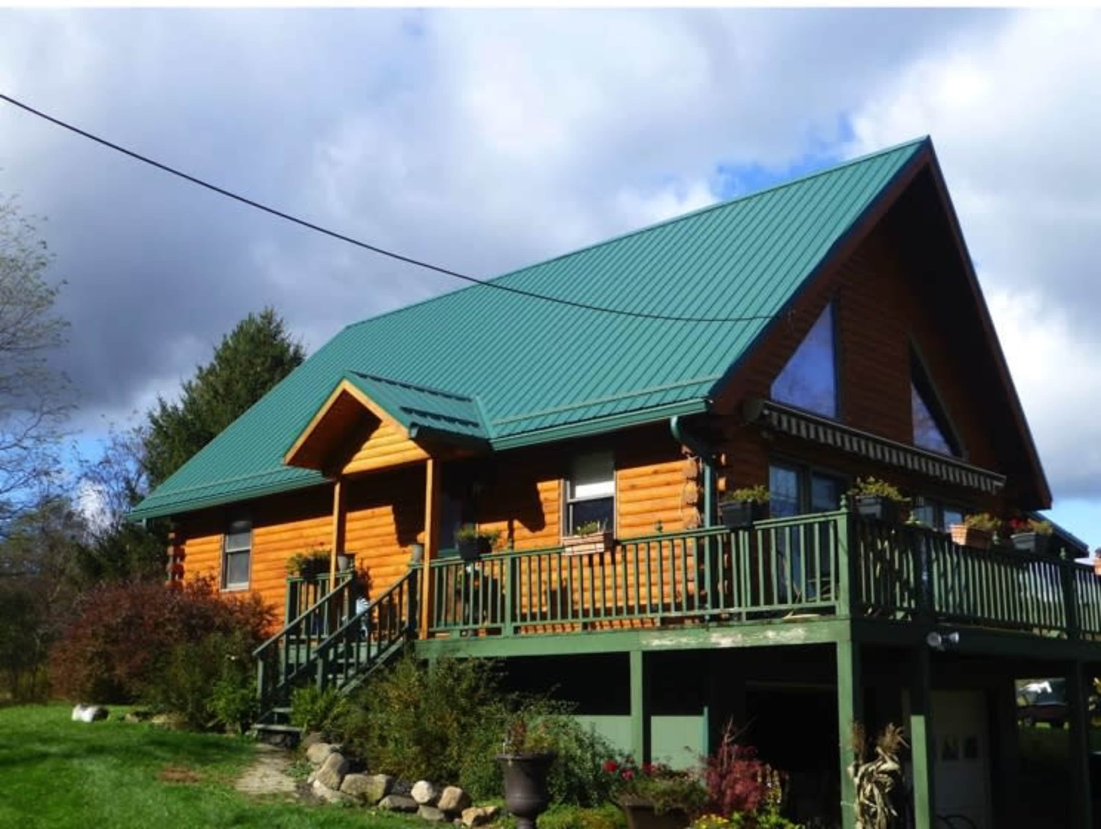 A log cabin with a green metal roof features a large deck and surrounding greenery.