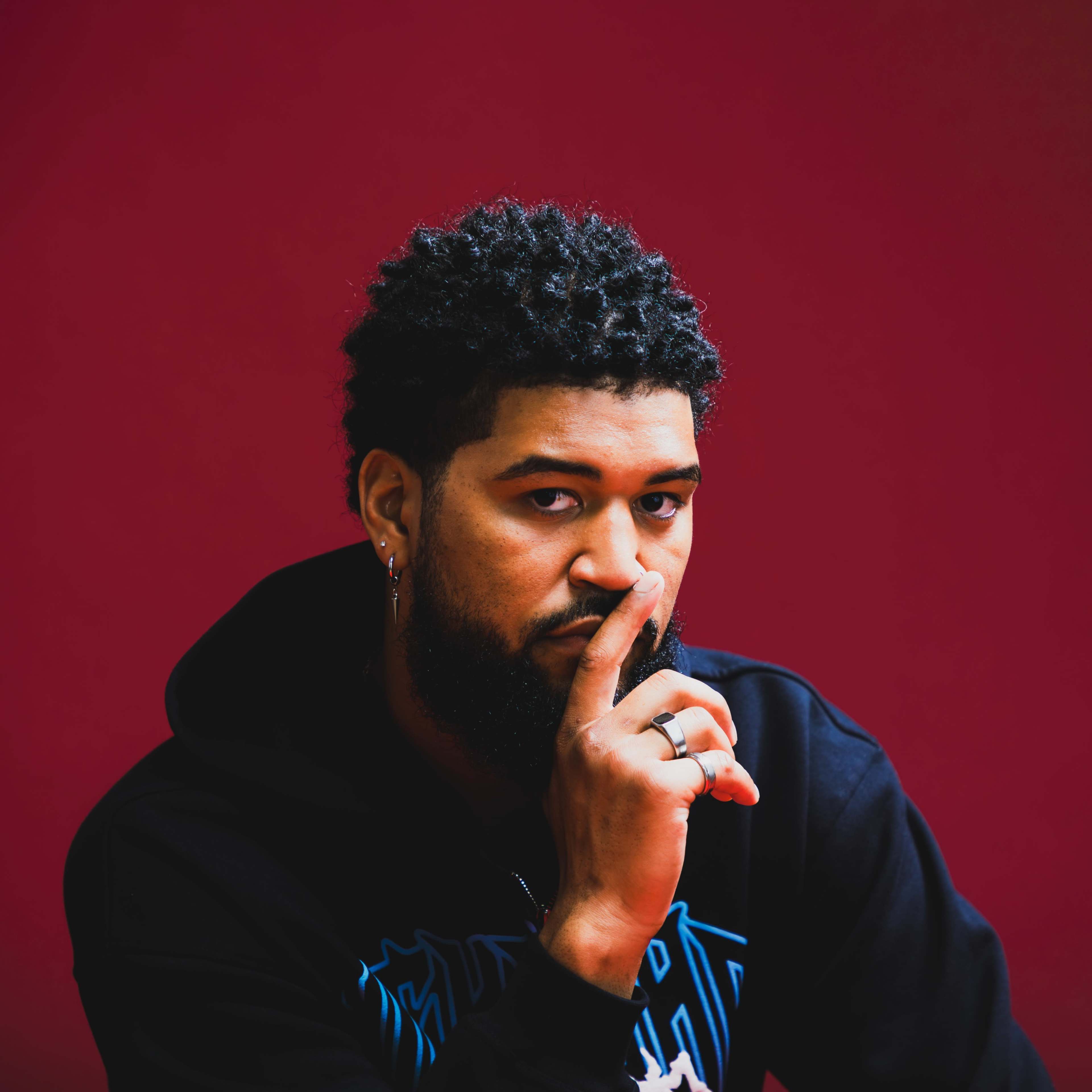 A man with curly hair and a beard is sitting against a red background, holding his finger to his lips in a "shh" gesture.