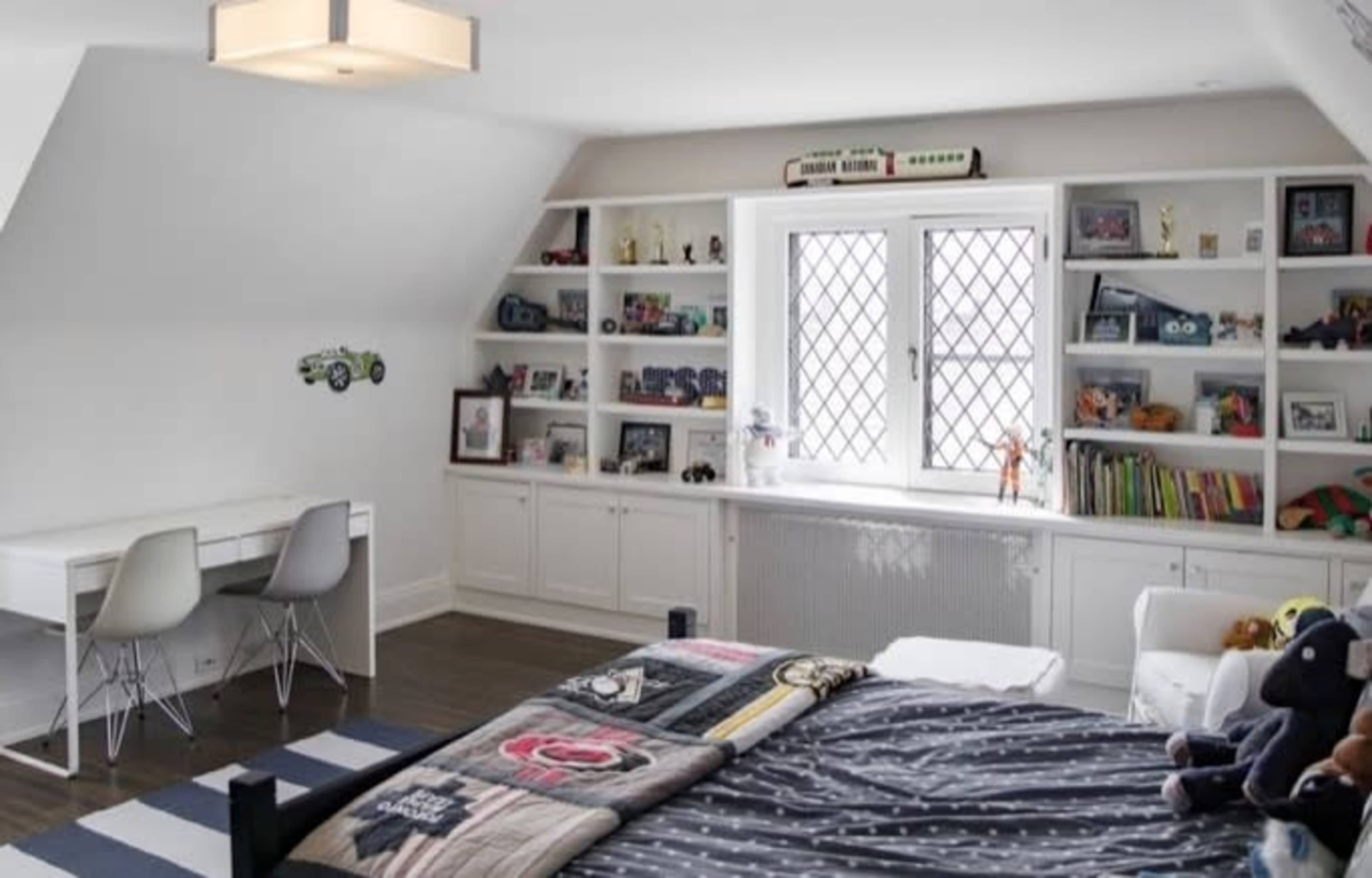 A child's bedroom featuring a bed with patterned bedding, a small desk with chairs, and shelves filled with books and toys under a window.