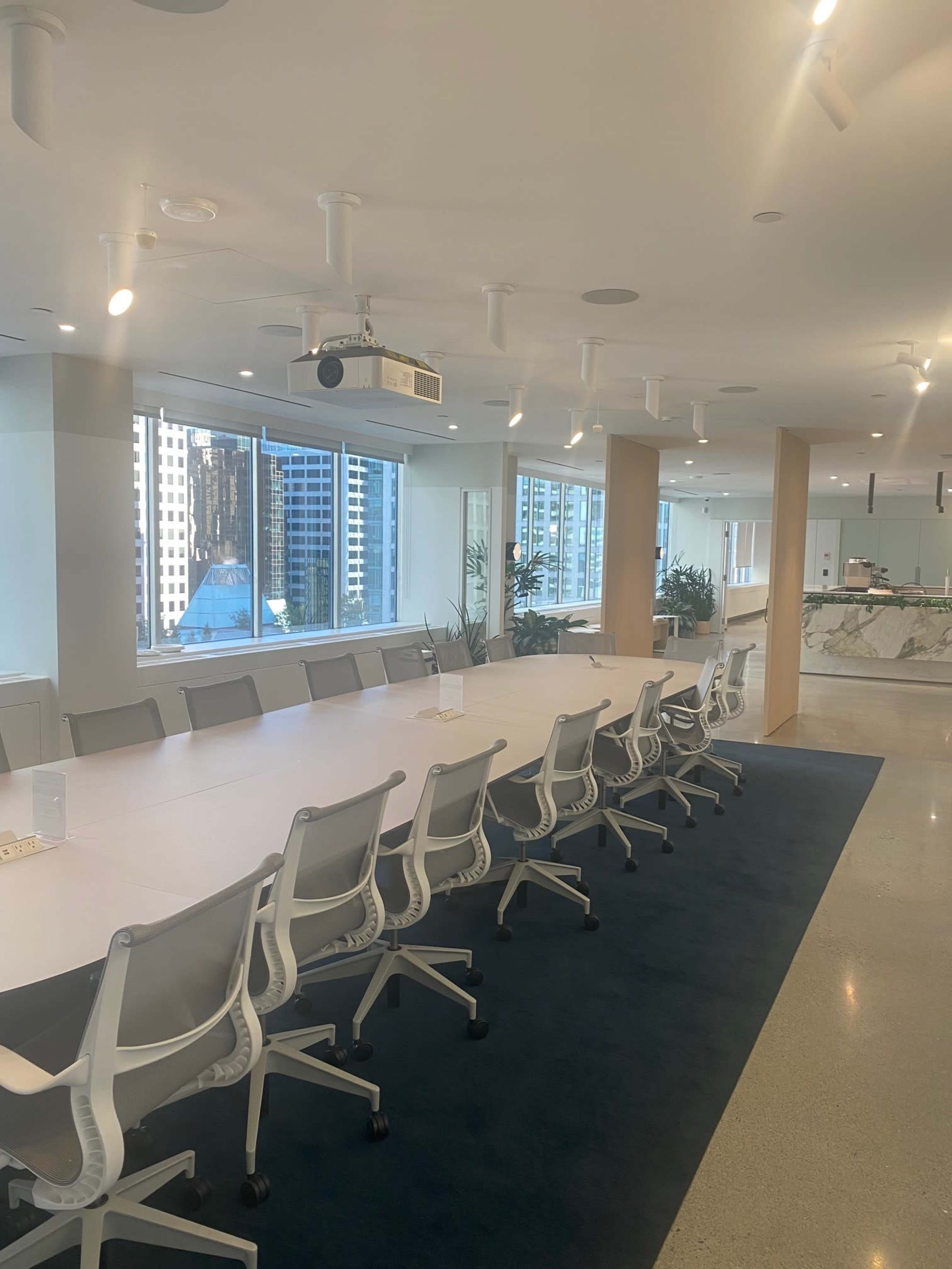 A large conference room features a long white table surrounded by gray chairs, with floor-to-ceiling windows overlooking a city skyline.