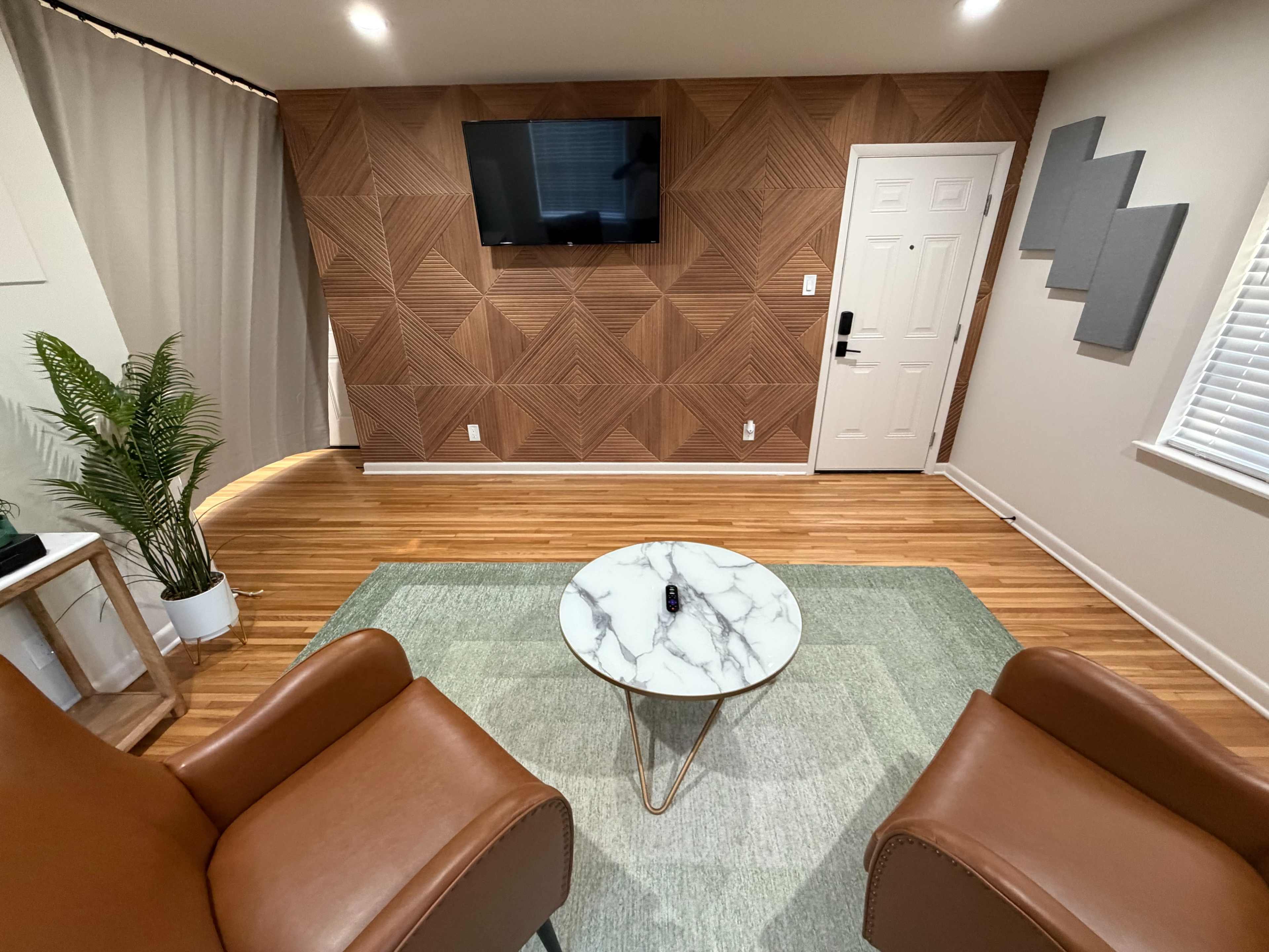 The image shows a modern living space with two brown armchairs facing a round marble coffee table on a green area rug, with a textured wood wall and a television mounted above a door.