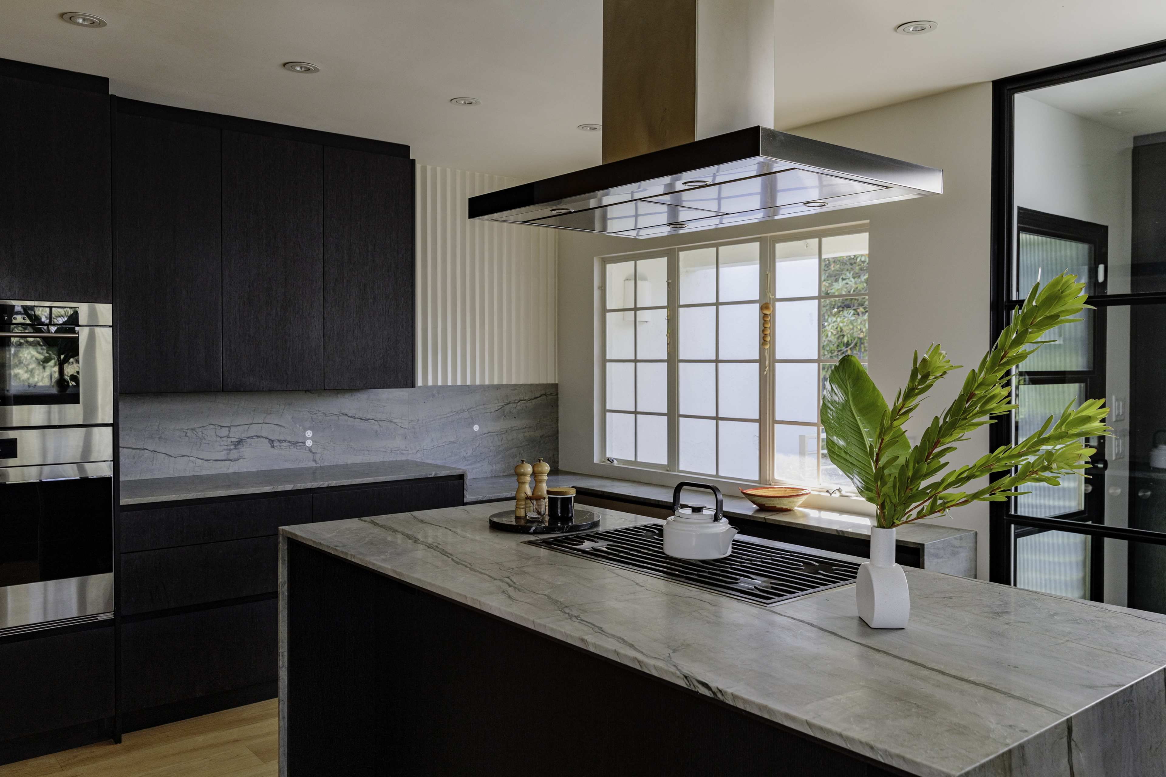 A modern kitchen features dark cabinetry, a central island with a stone countertop, and a stainless steel range hood above a stovetop.