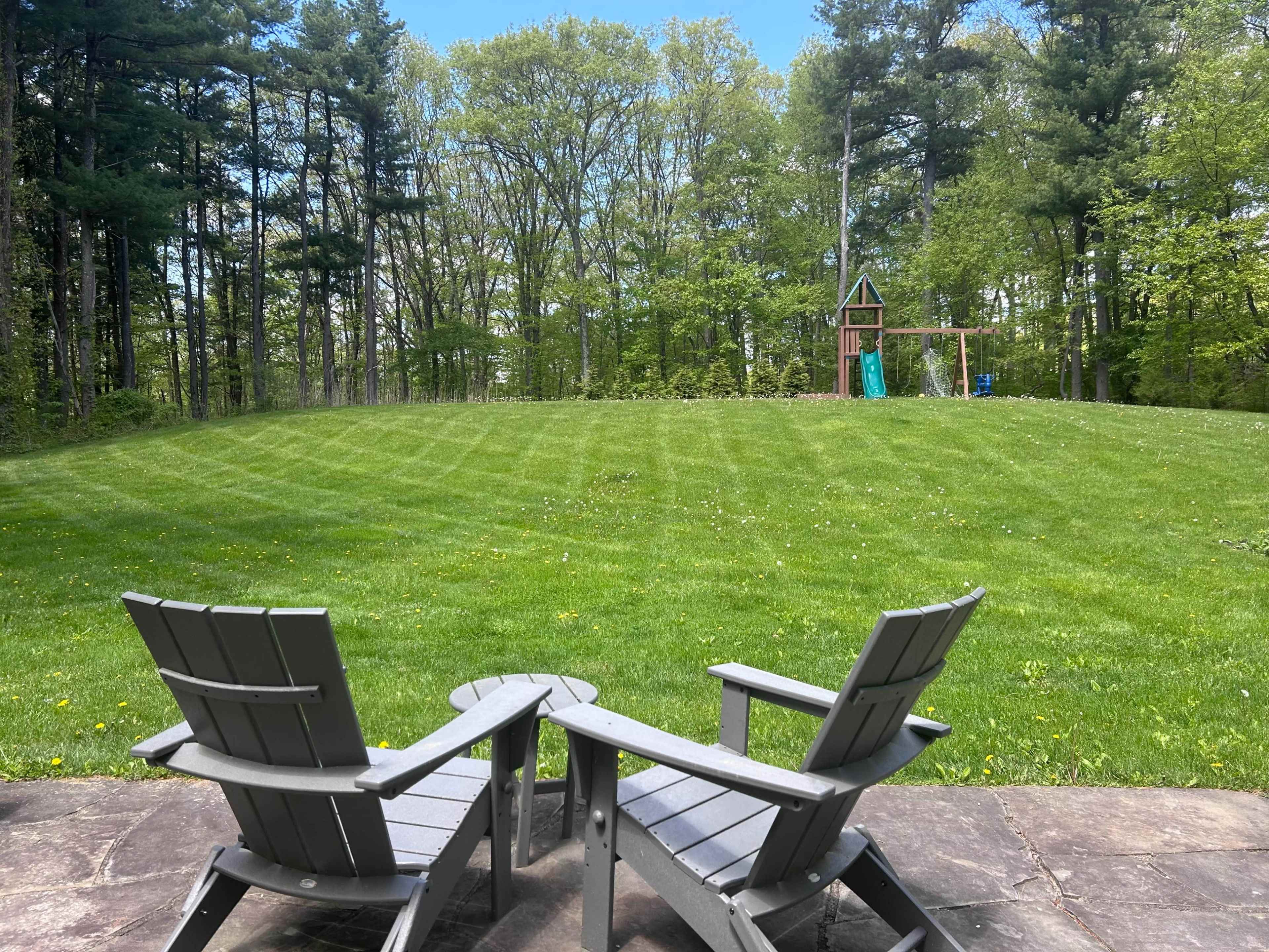 Two gray Adirondack chairs face a lush green lawn with a playground set in the background, surrounded by trees under a clear blue sky.