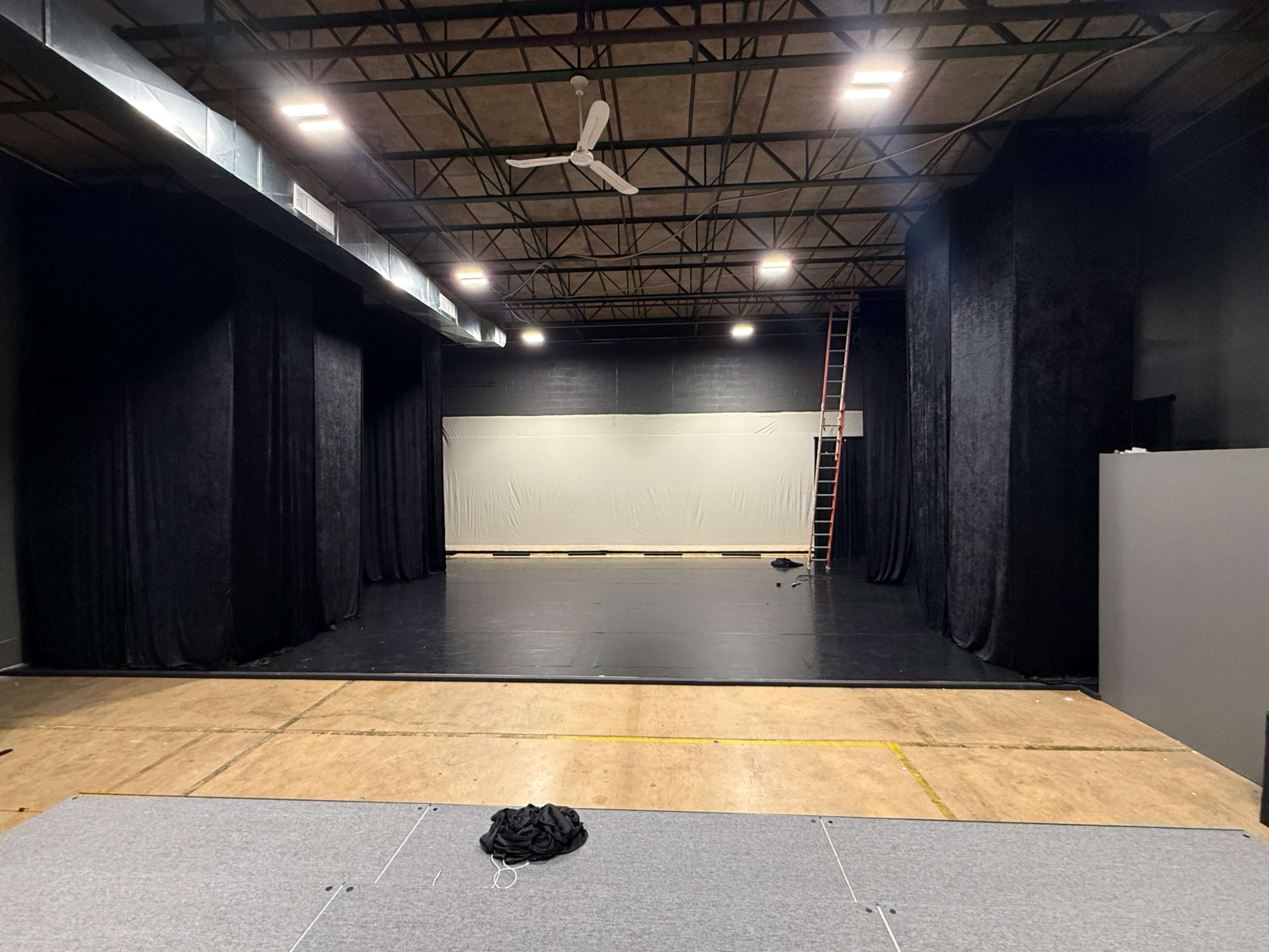 The image shows a large, empty performance space with black curtains on either side, a ladder against the wall, and a gray floor covering in the foreground.