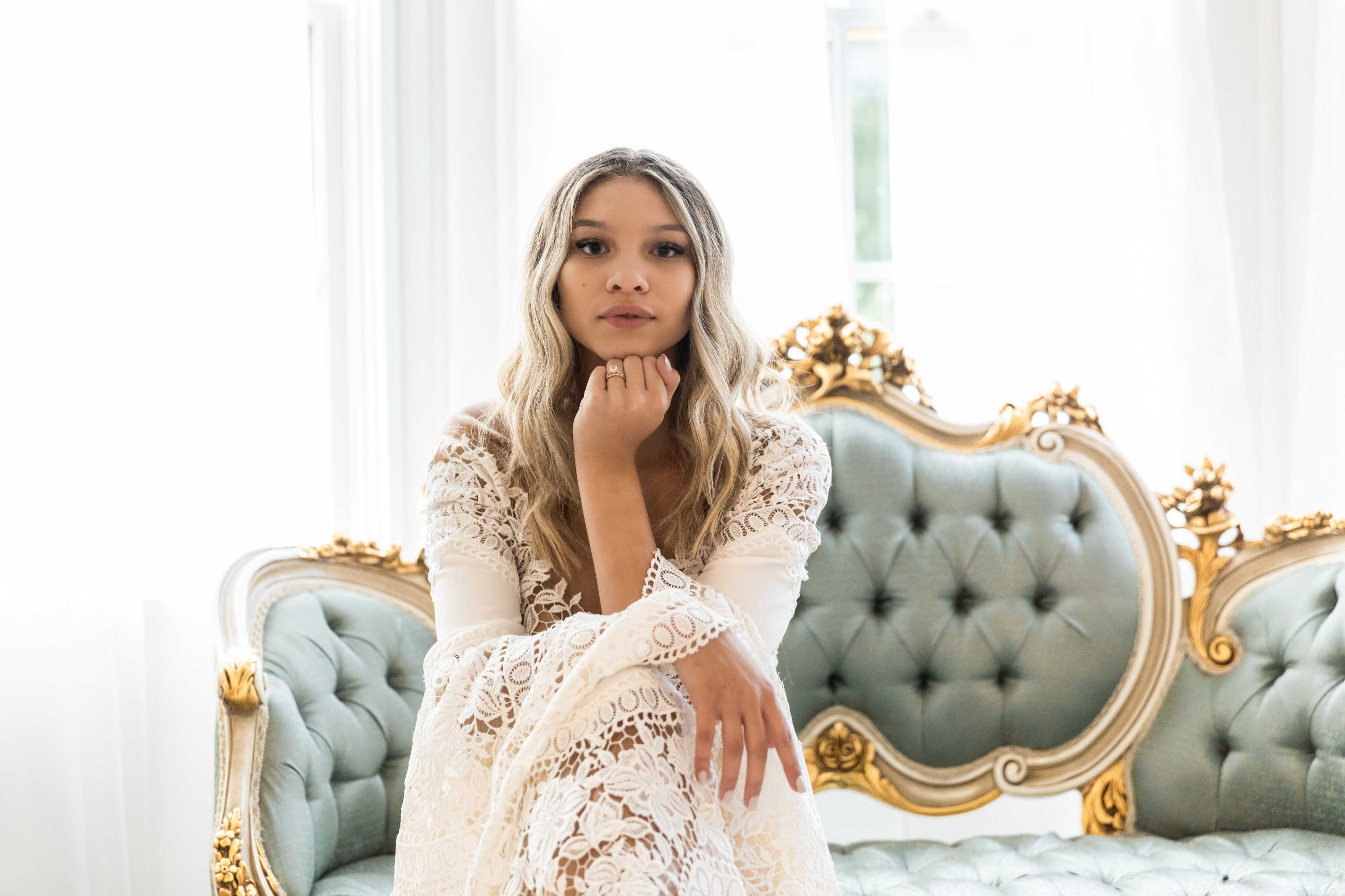 A woman in a white lace dress sits on an ornate couch with light streaming in from behind her.