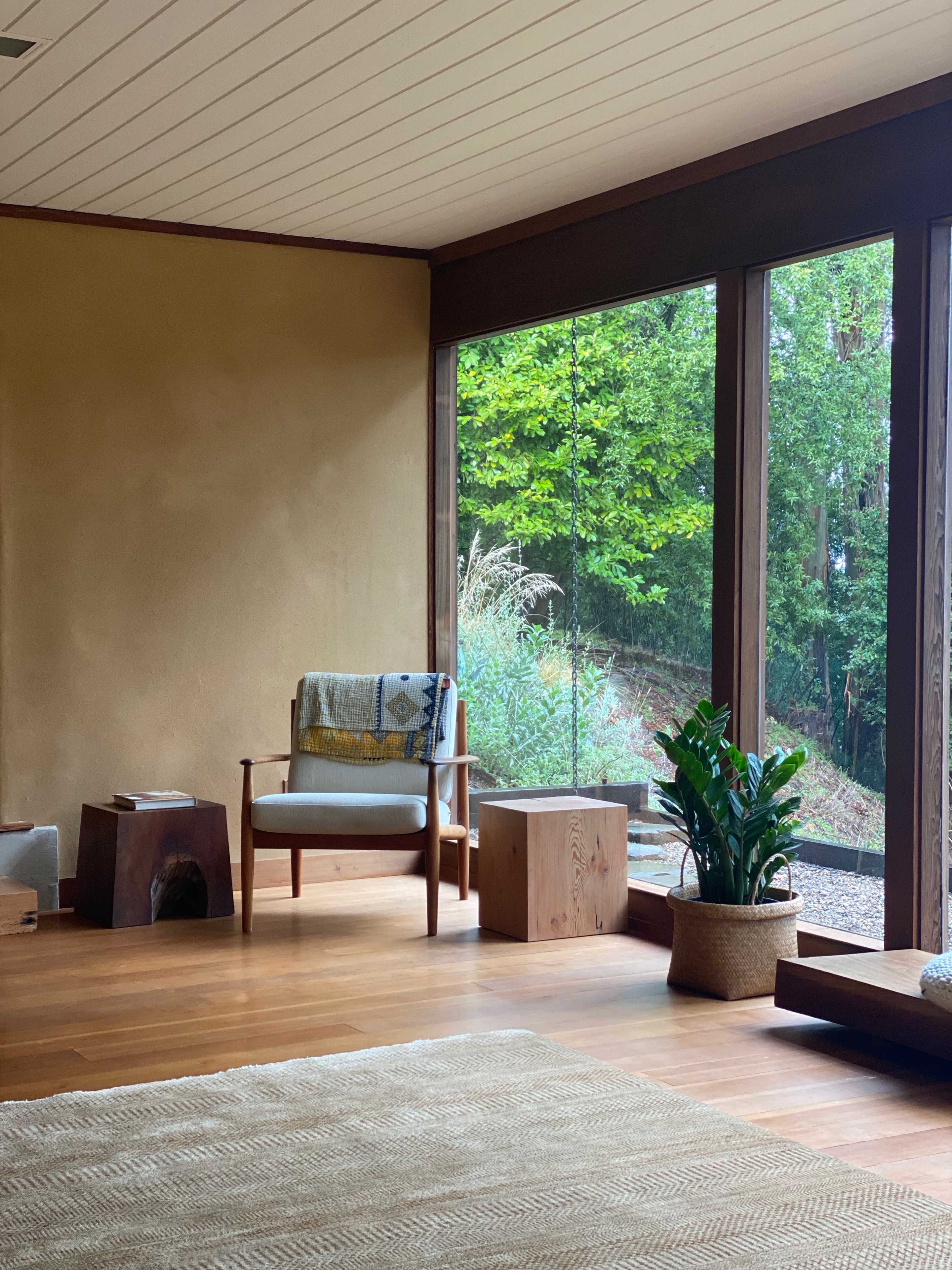 A minimalist living space features a wooden chair with a blanket, a potted plant, and large glass windows overlooking a green landscape.