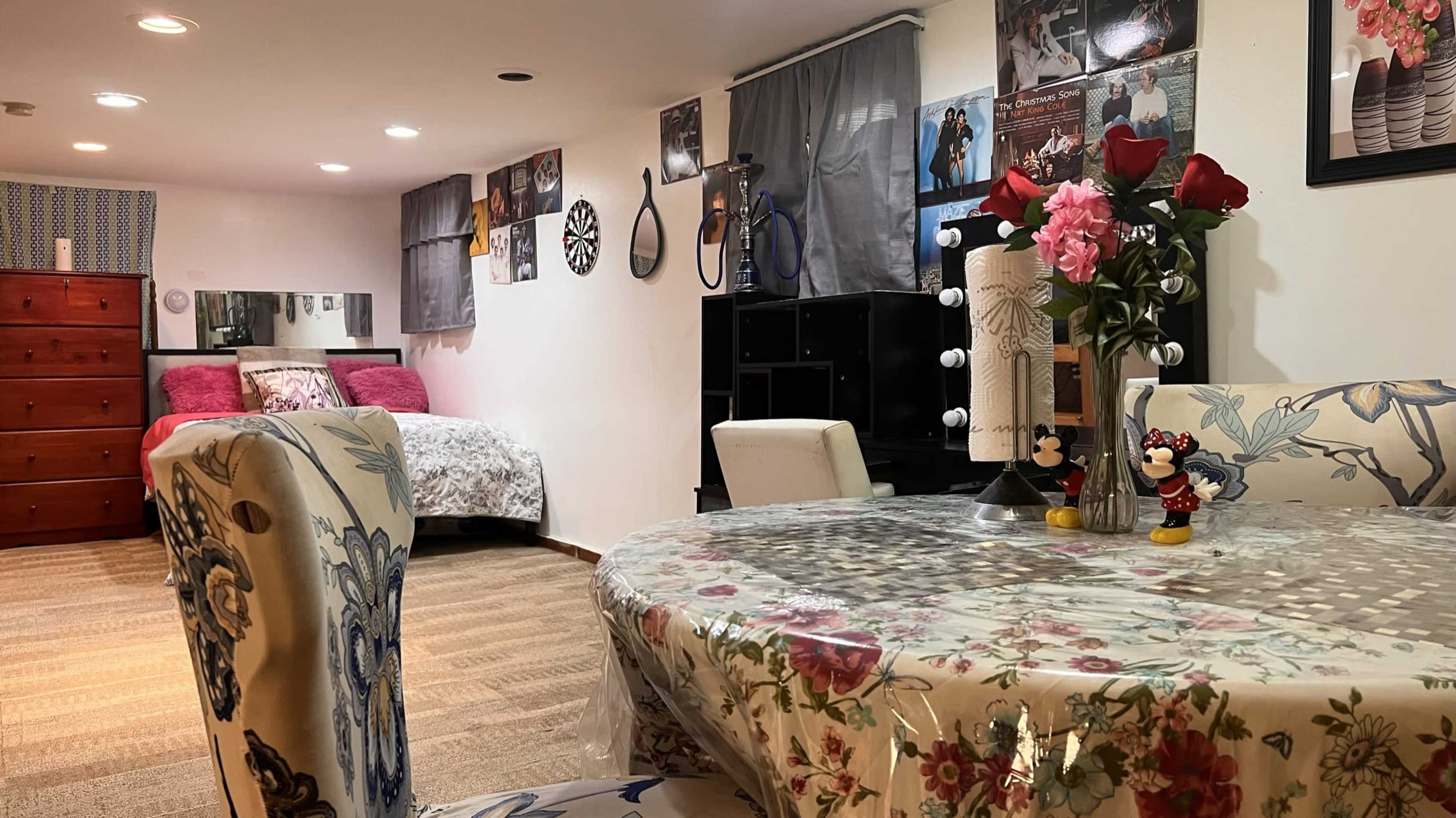 A cozy room featuring a dining table with floral decor, a bed with pink bedding in the background, and various decorations on the walls.