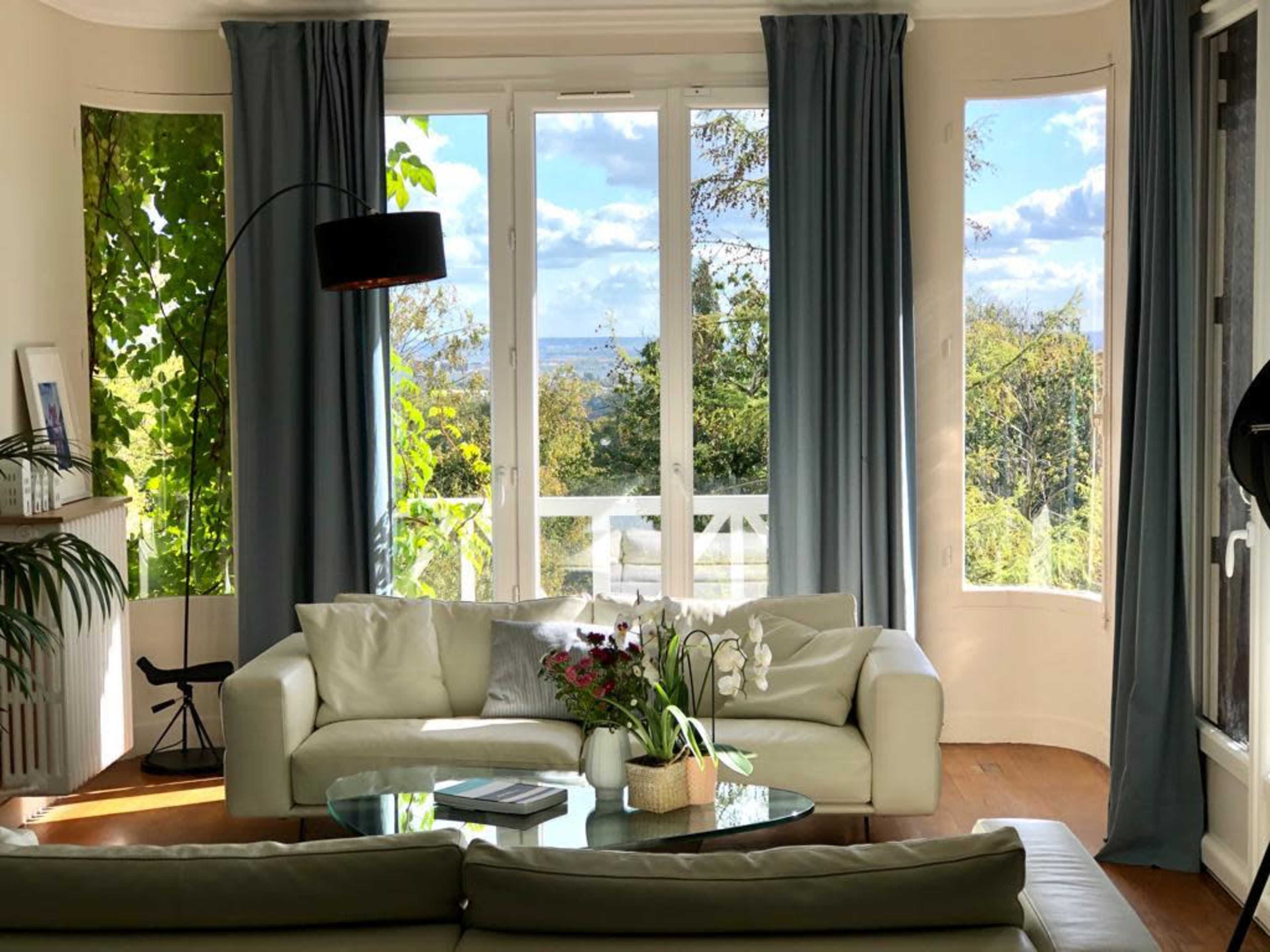 A bright living room features large windows with blue curtains, a white leather sofa, and a glass coffee table adorned with flowers.