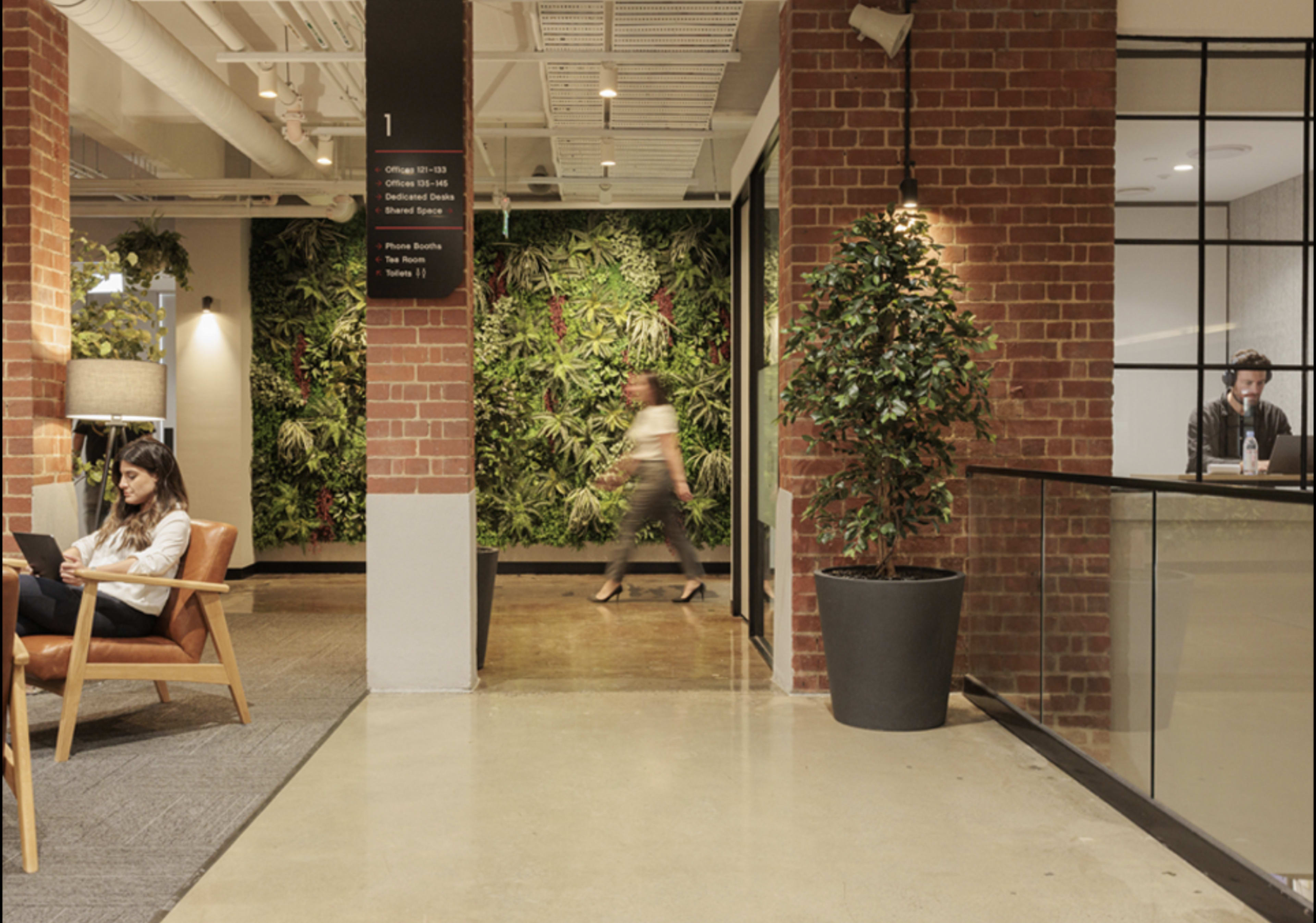 A modern office space featuring a green wall, a seating area with a woman using a phone, and a man working at a reception desk in the background.