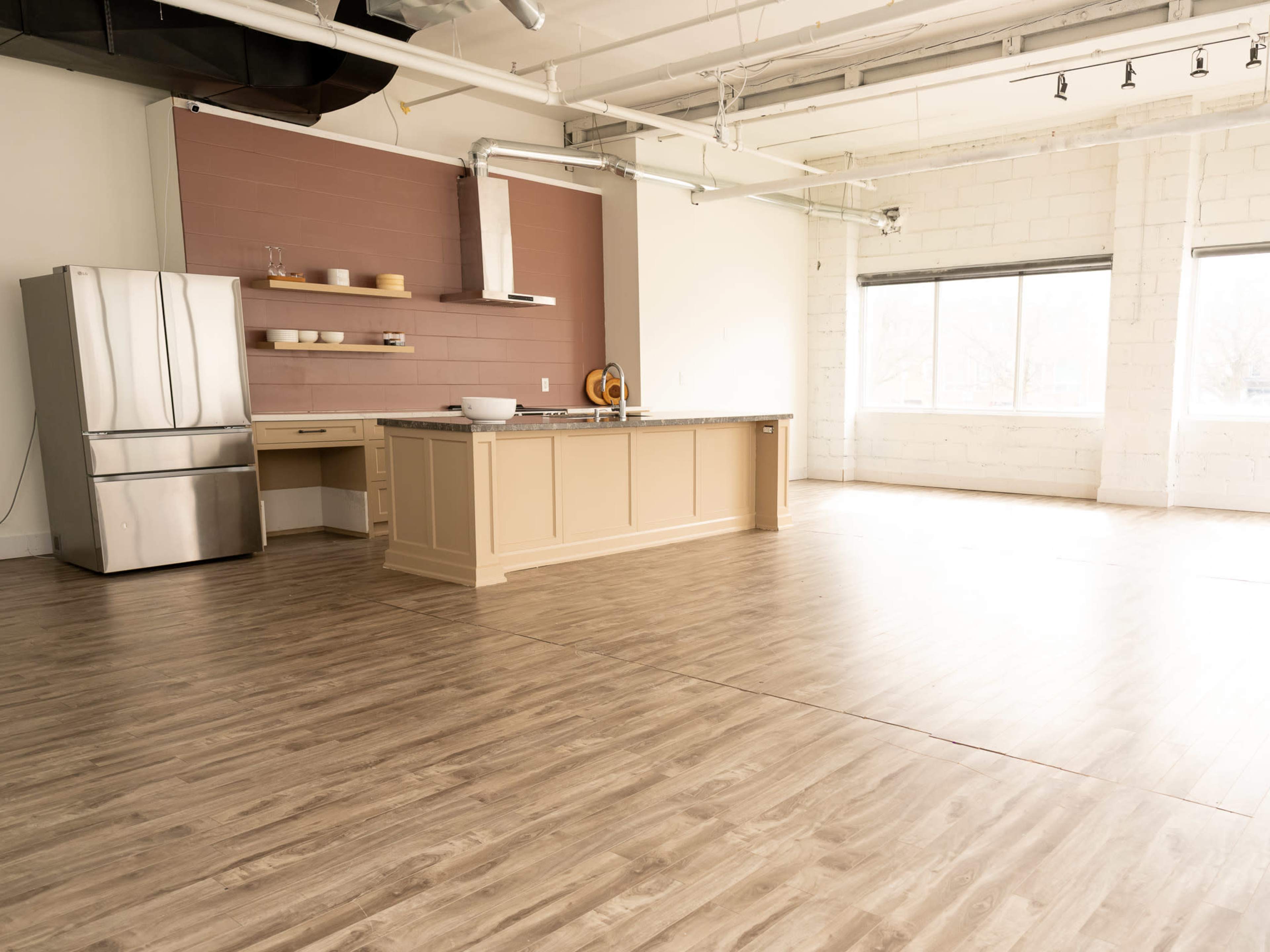 An empty modern kitchen space features a large island, stainless steel refrigerator, and light wood flooring.
