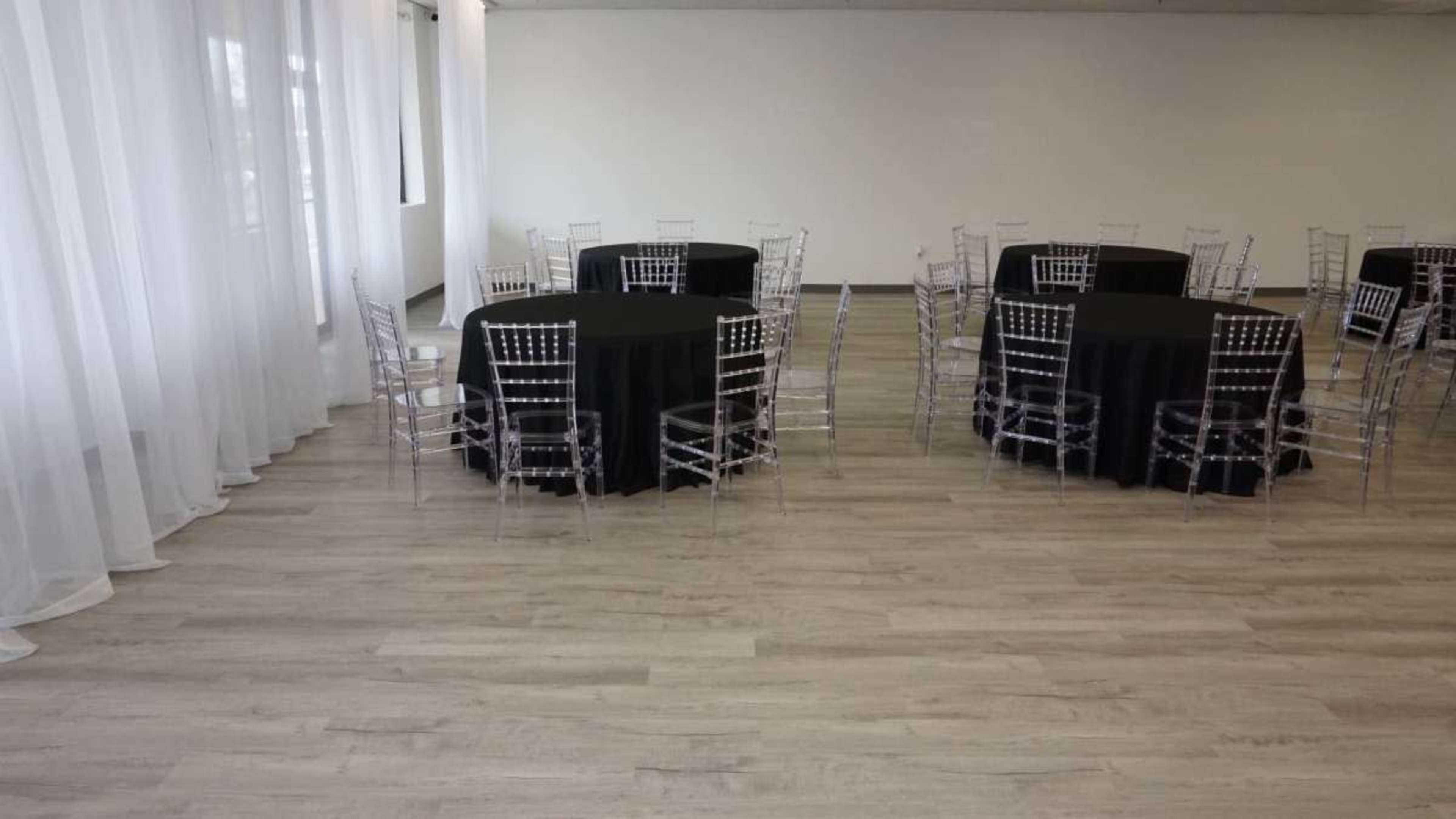 The image shows a spacious indoor venue with several round tables covered in black tablecloths, surrounded by transparent chairs, and large sheer curtains along one wall.