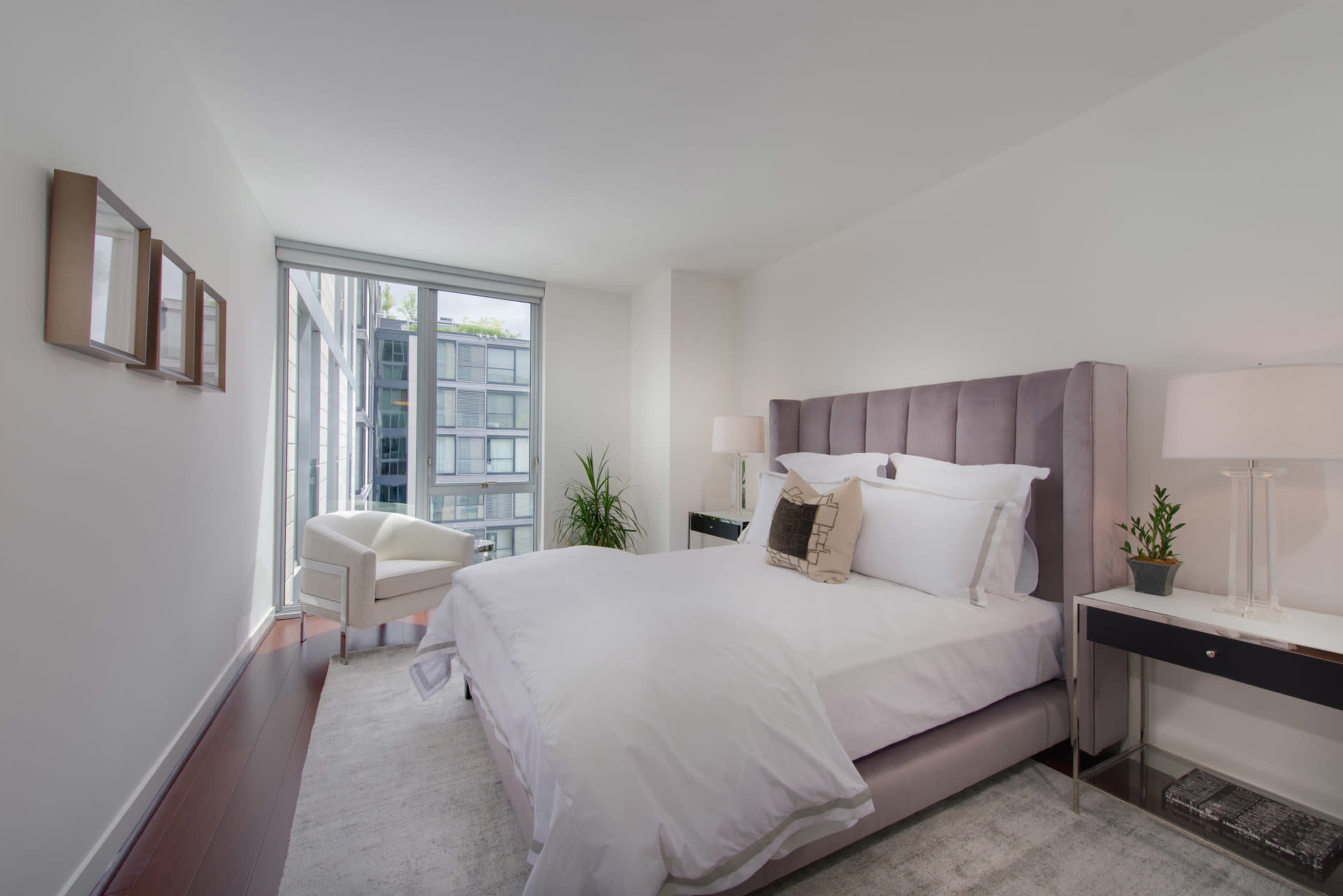 The image shows a modern bedroom featuring a large bed, two bedside lamps, a chair by the window, and minimal decor.