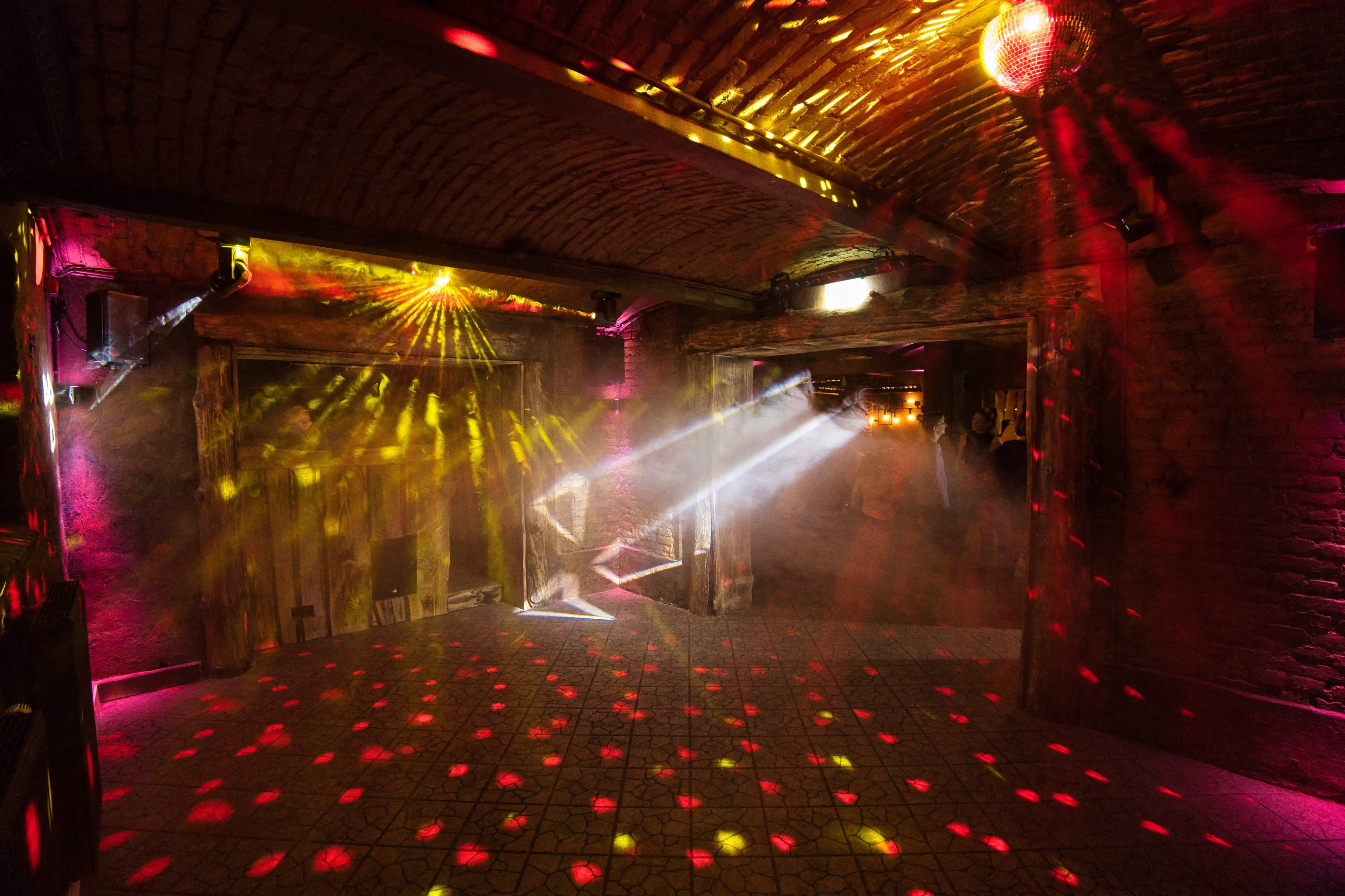 The interior of a dimly lit venue features colorful light beams and a disco ball casting patterns across the floor and walls.