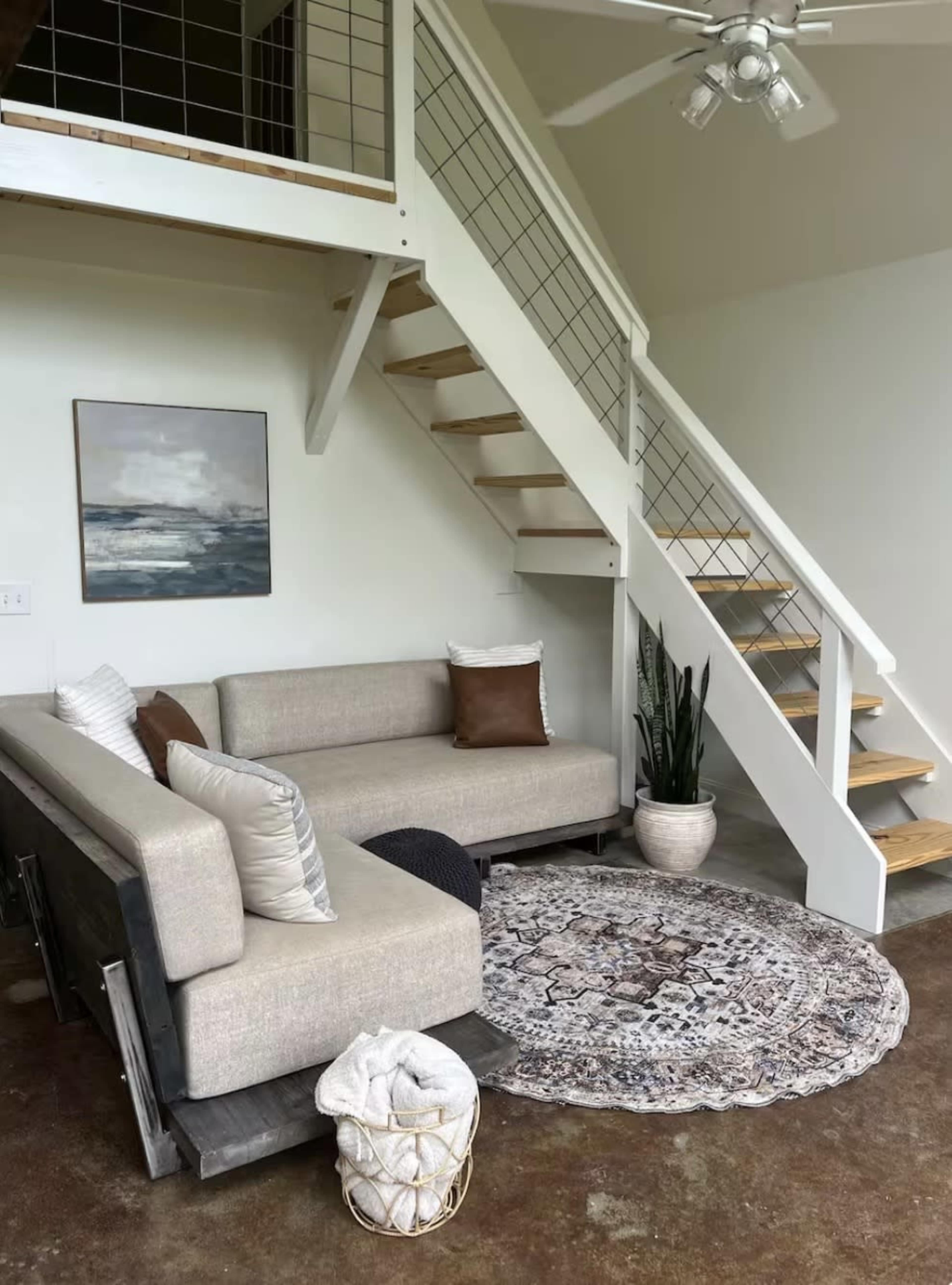 The image shows a modern living room with a beige sectional sofa, a round area rug, and a staircase leading to a lofted space.