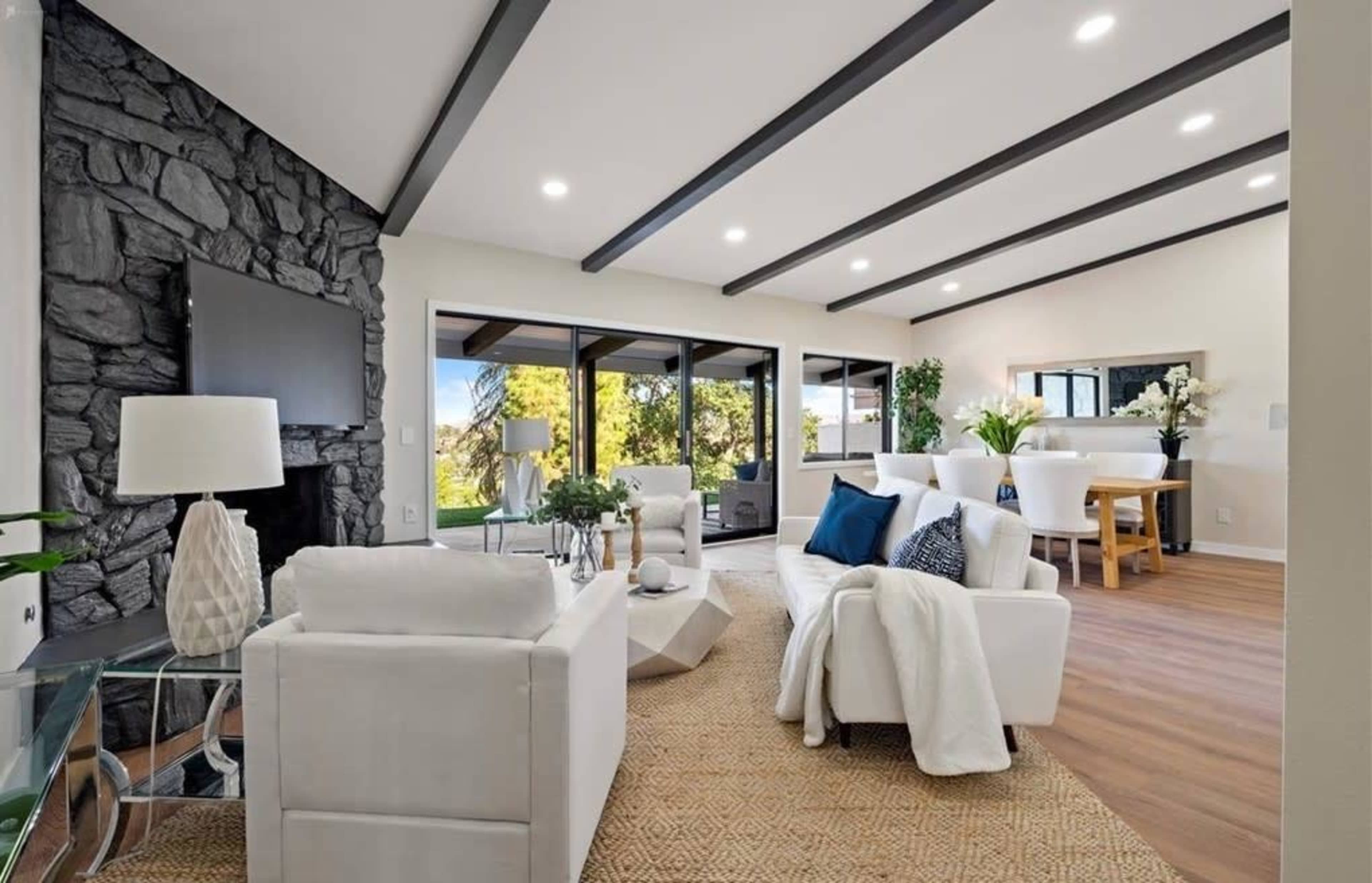 A modern living room features a stone accent wall, white furniture, large windows, and a dining area with a round table and plants.