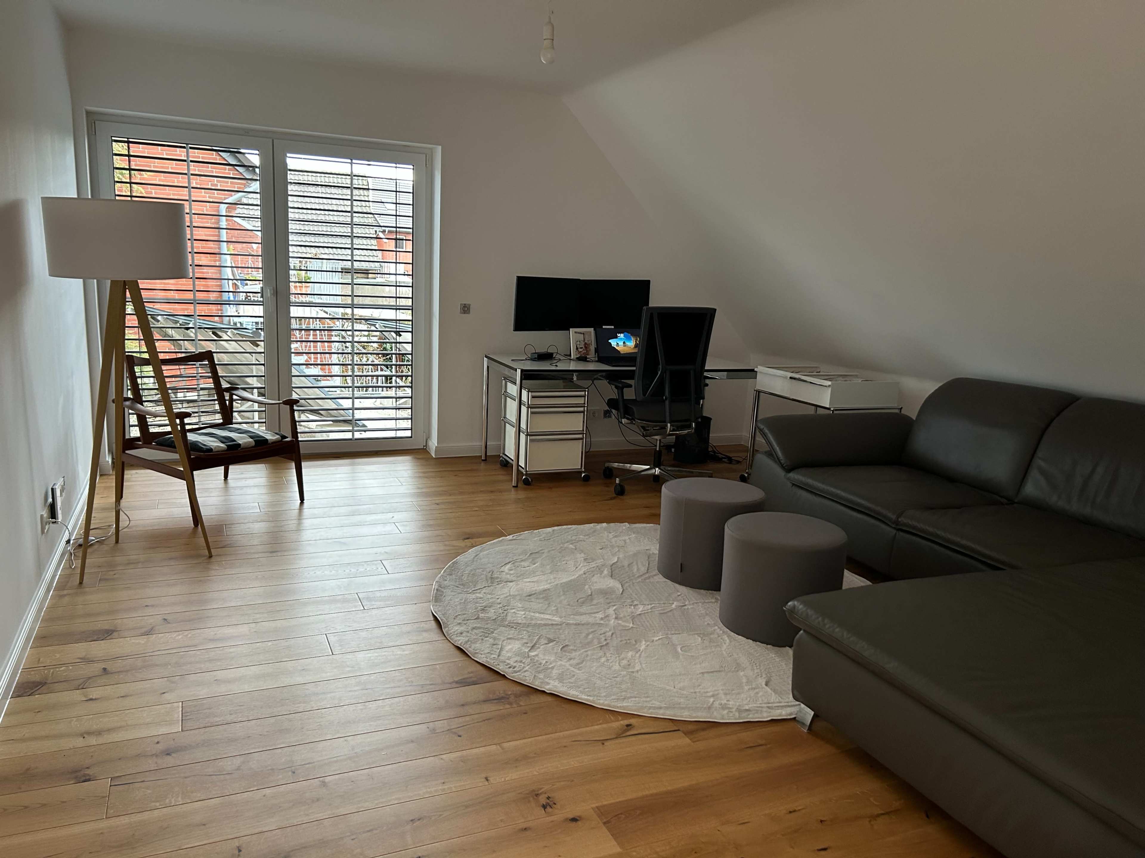 The image shows a minimalist living space featuring a leather sectional sofa, a round rug, two gray stools, a desk with a computer, and a chair, all set against a backdrop of large windows.