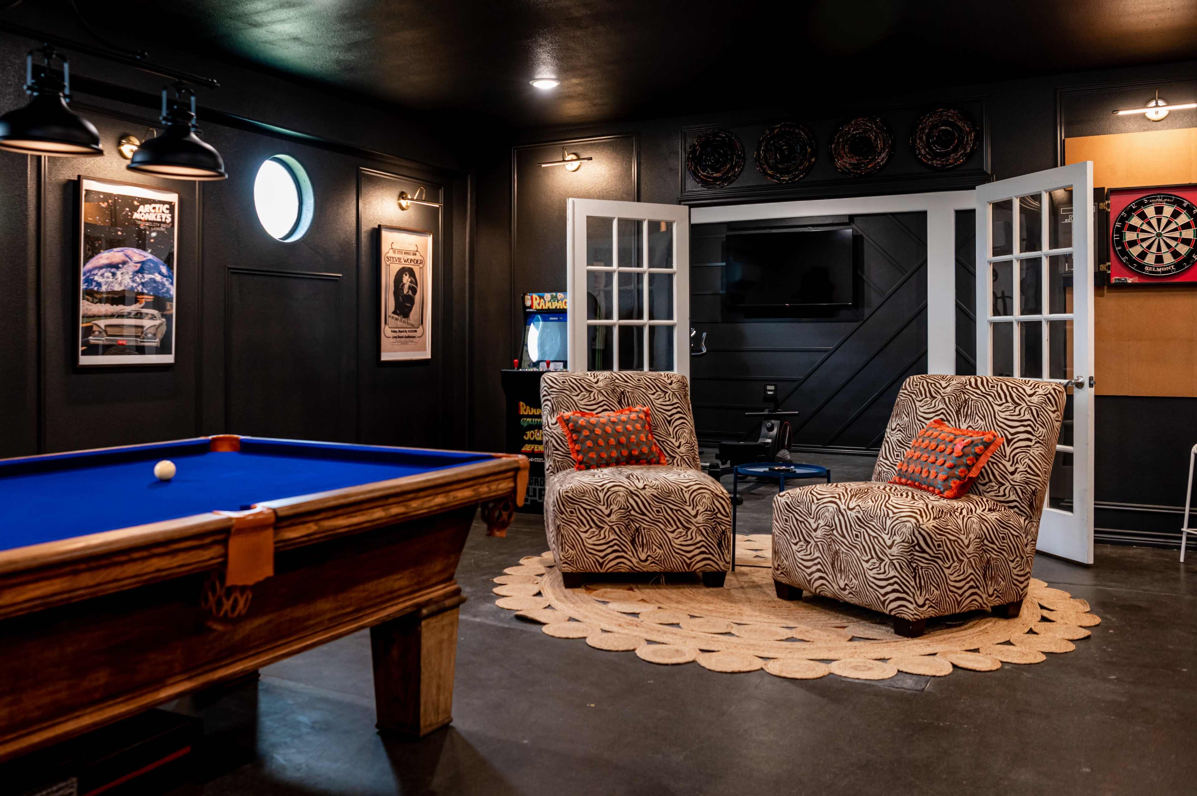 A game room featuring a blue pool table, two patterned armchairs, a round rug, dartboard, and arcade machine, with double doors leading to another space.