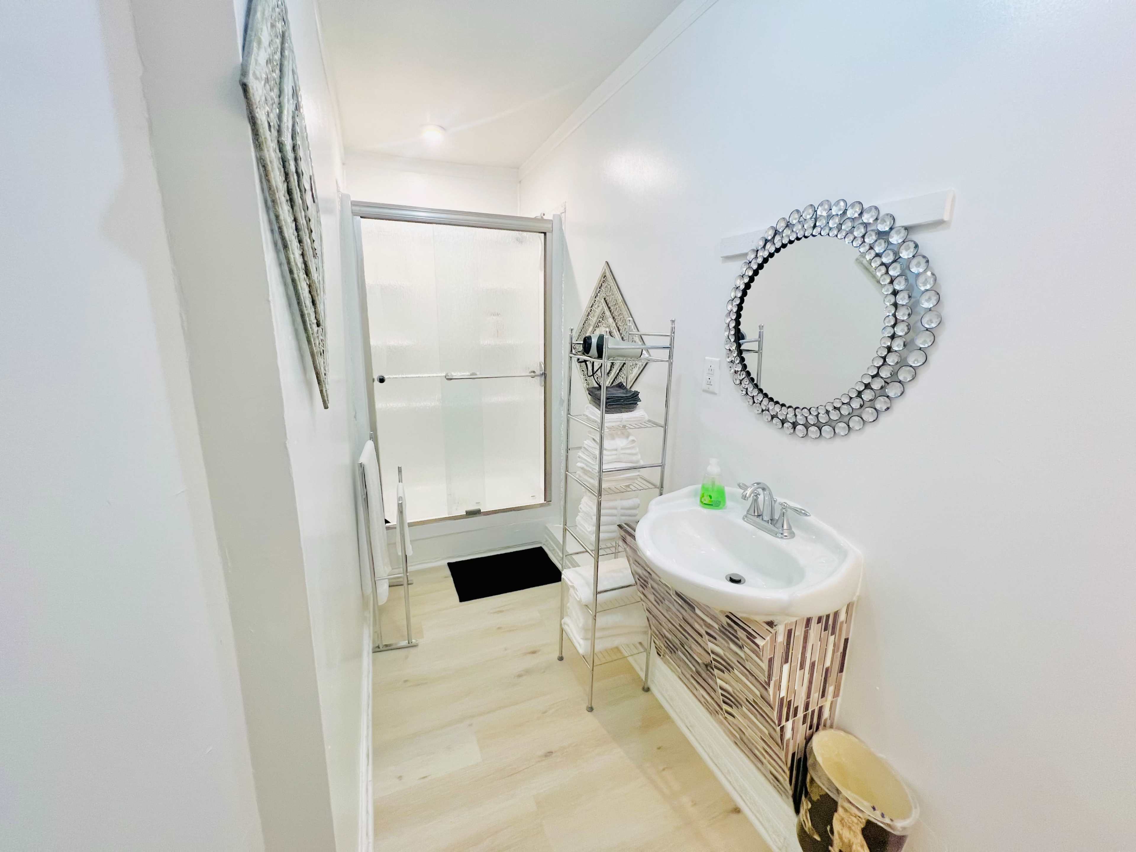 A bathroom with a circular mirror, a sink, shelves of white towels, and a glass shower enclosure.