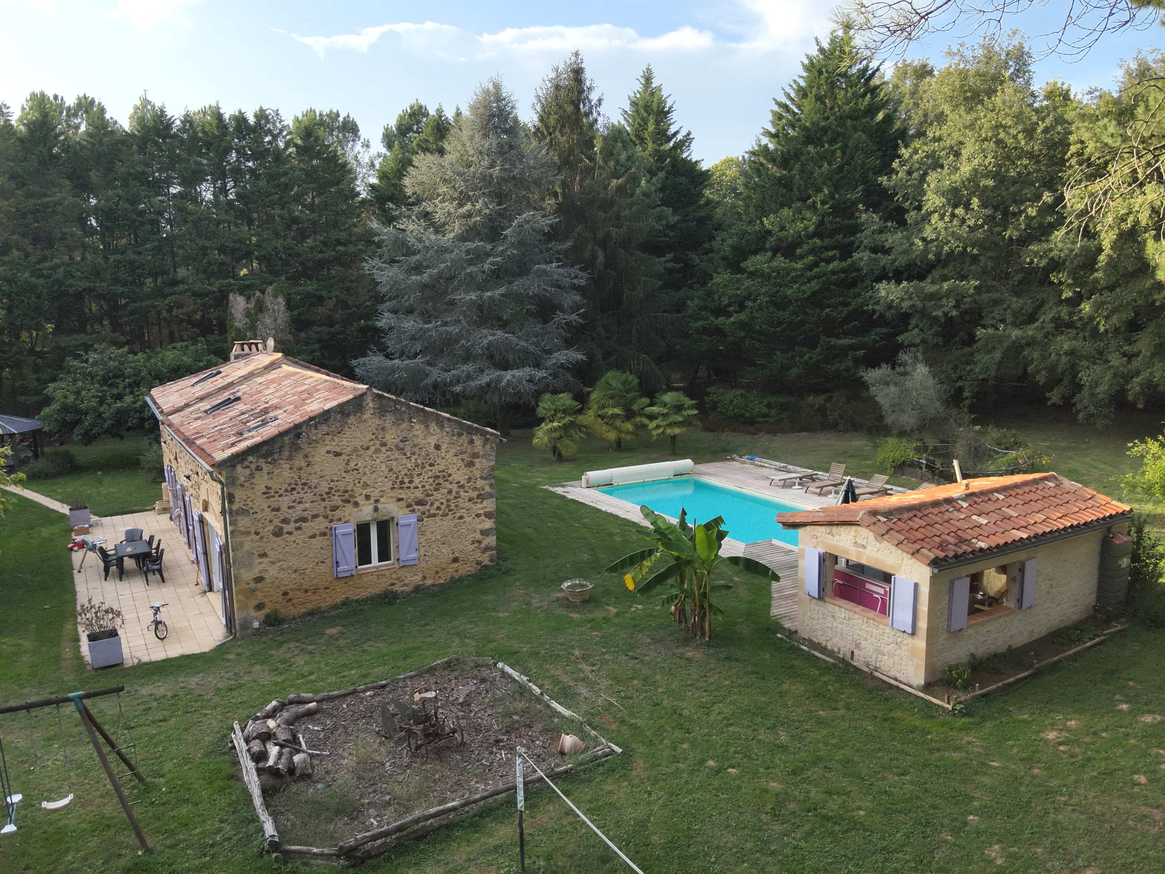 The image shows a rural property with two stone houses, a swimming pool, and a landscaped garden surrounded by trees.