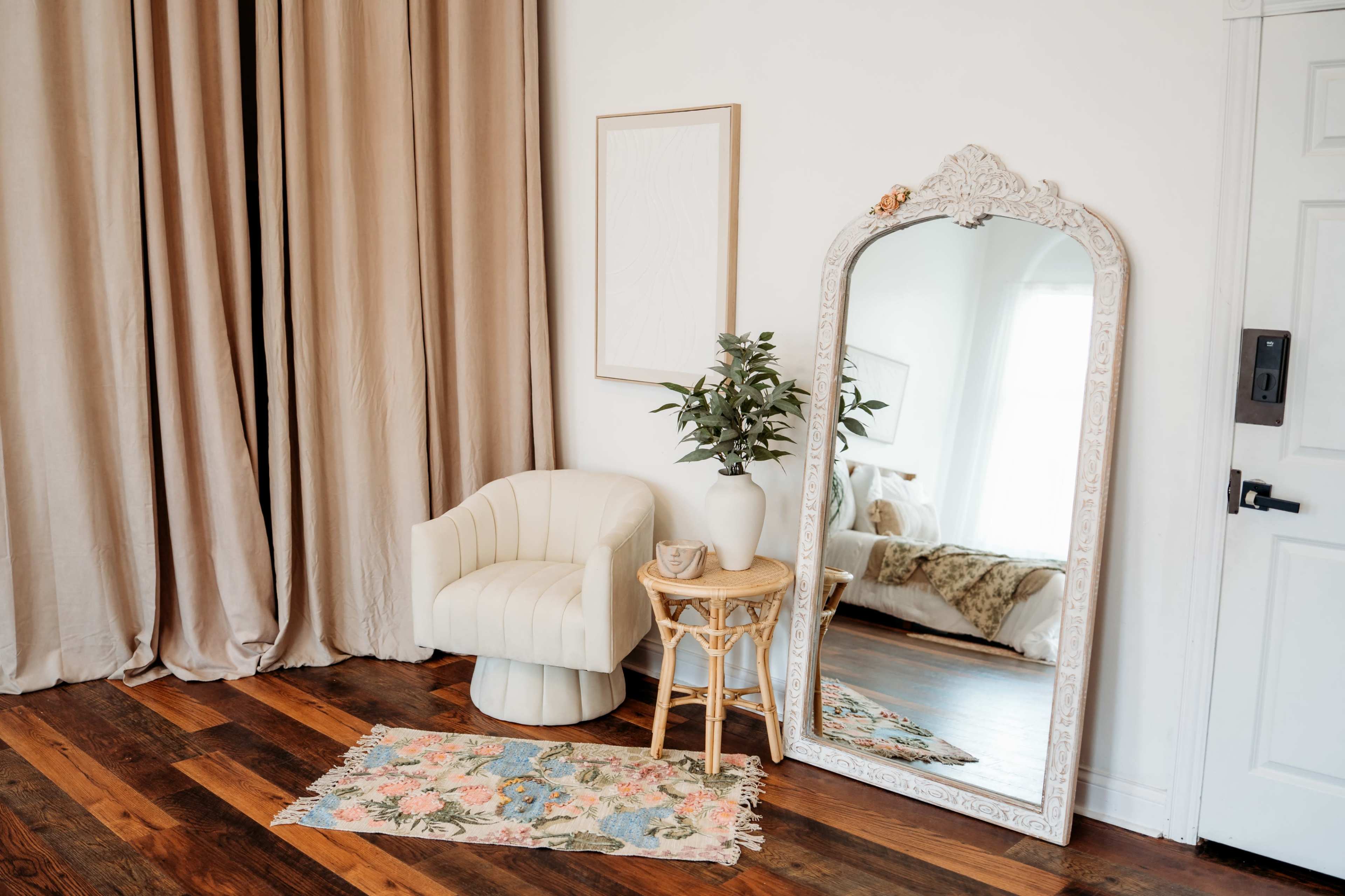 The image features a cozy room with a large decorative mirror, a light-colored chair, a small side table, and a floral rug set against a backdrop of beige curtains and wooden flooring.