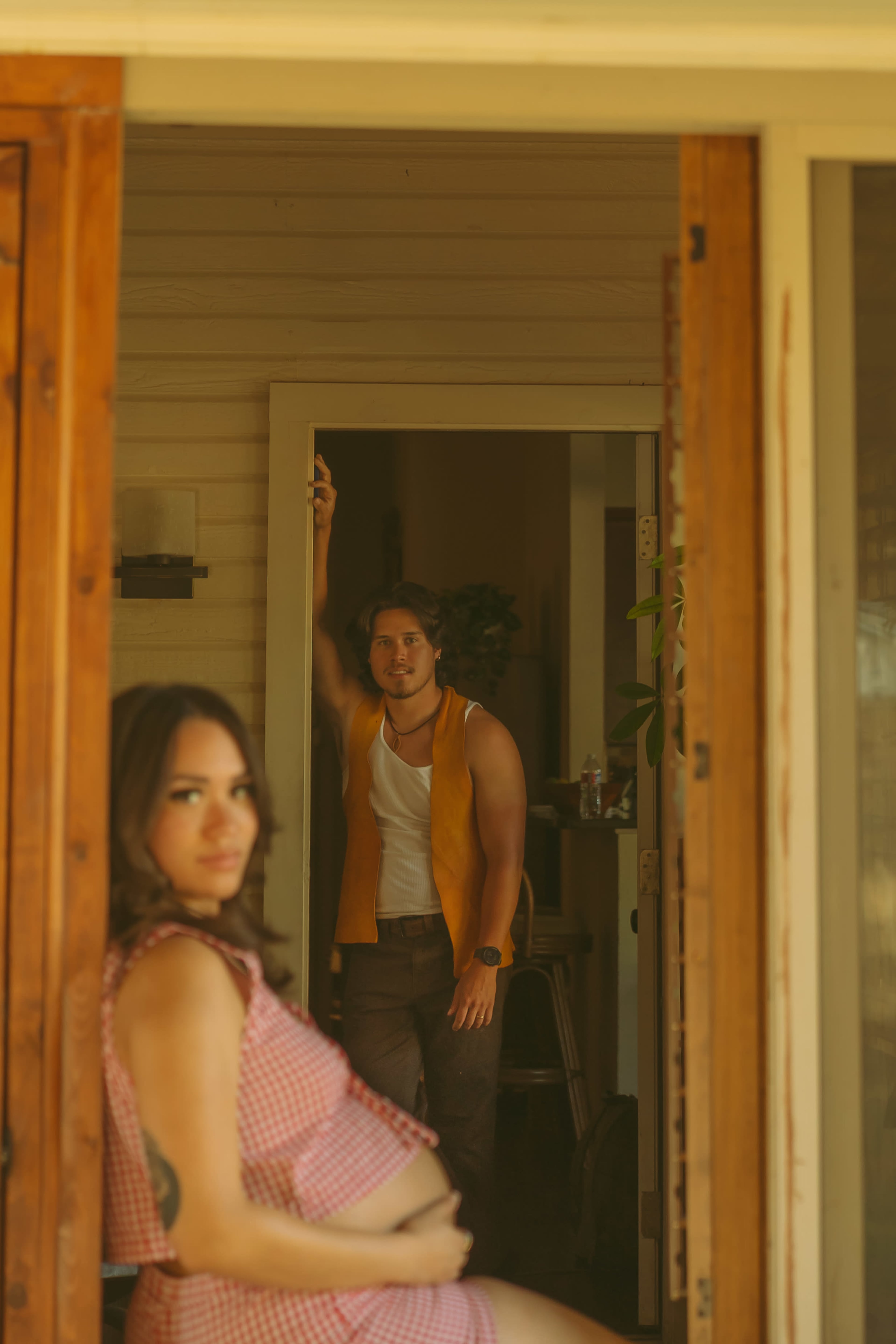 A woman in a checked outfit sits in the foreground while a man in a sleeveless shirt stands in an open doorway behind her.