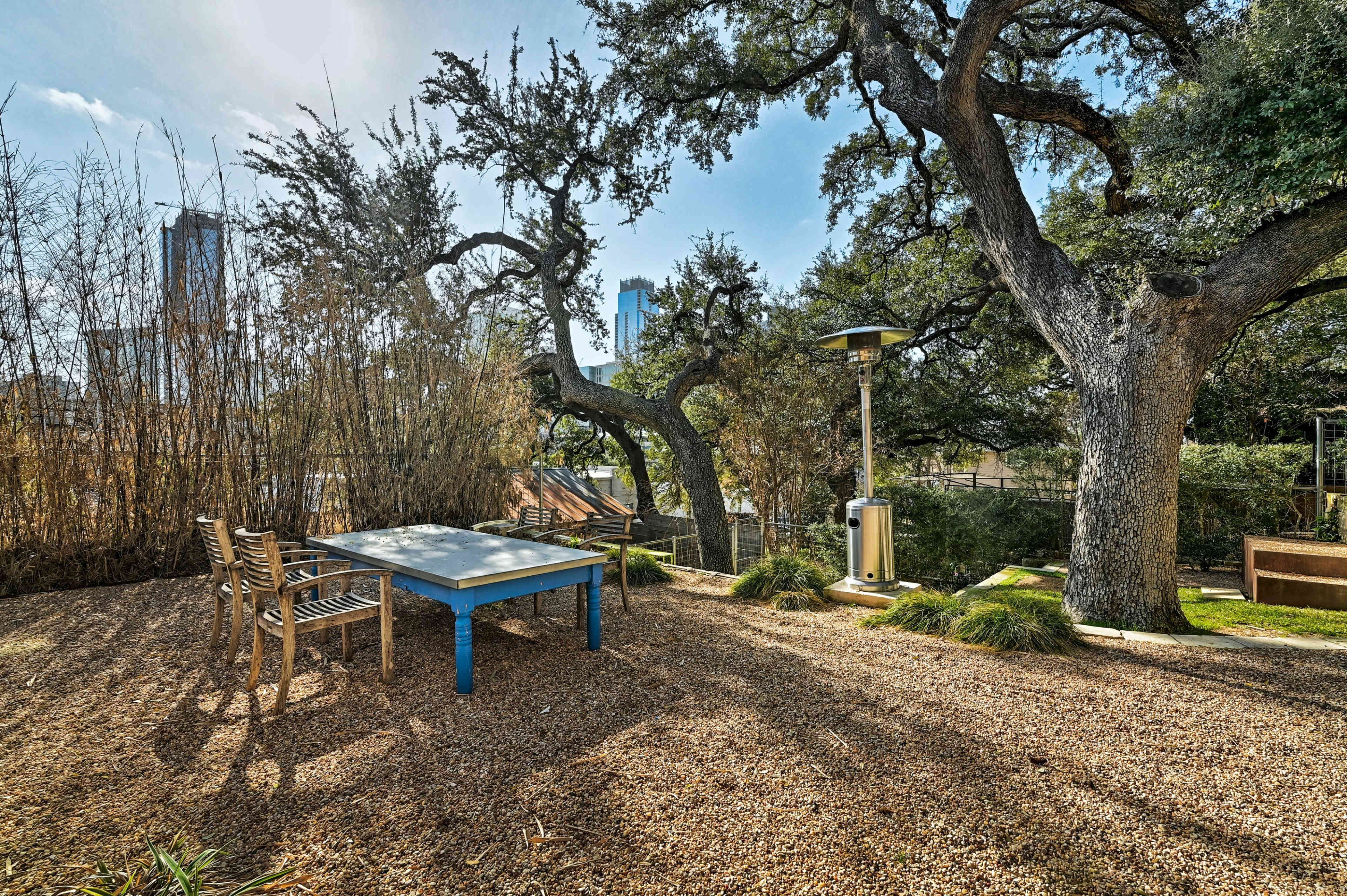 A table with chairs is set up on a gravel area surrounded by trees and tall buildings in the background.