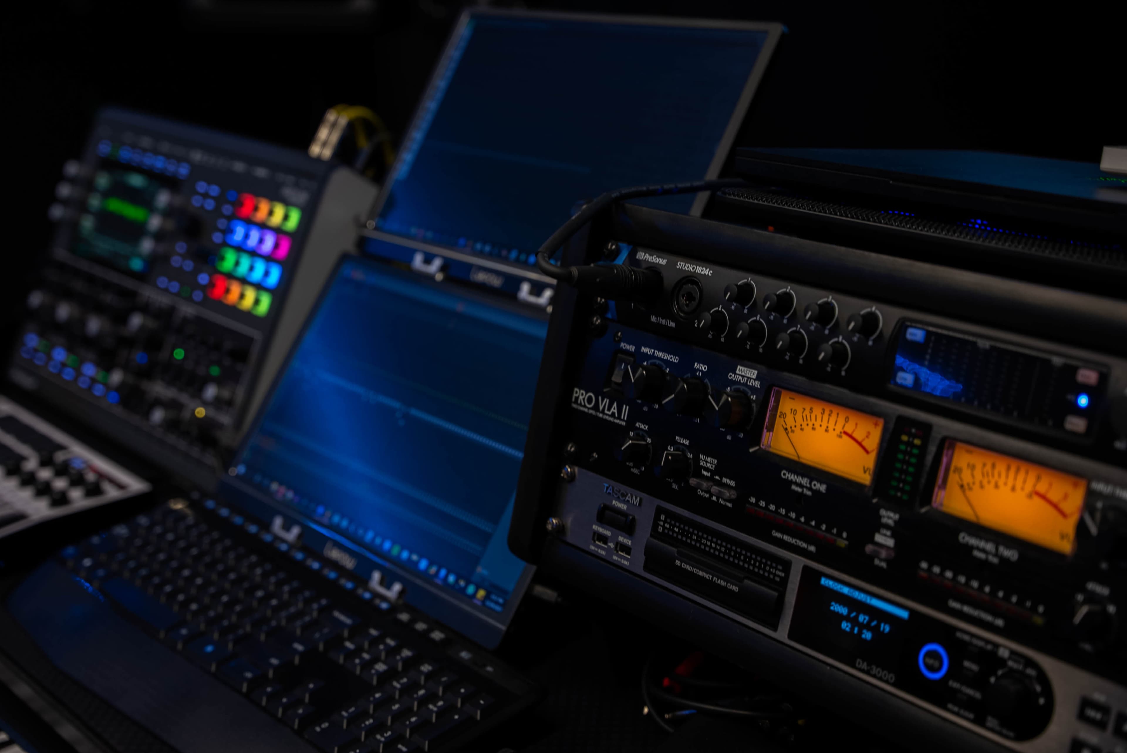 The image shows a control room setup featuring multiple monitors and audio equipment with various lights and meters.