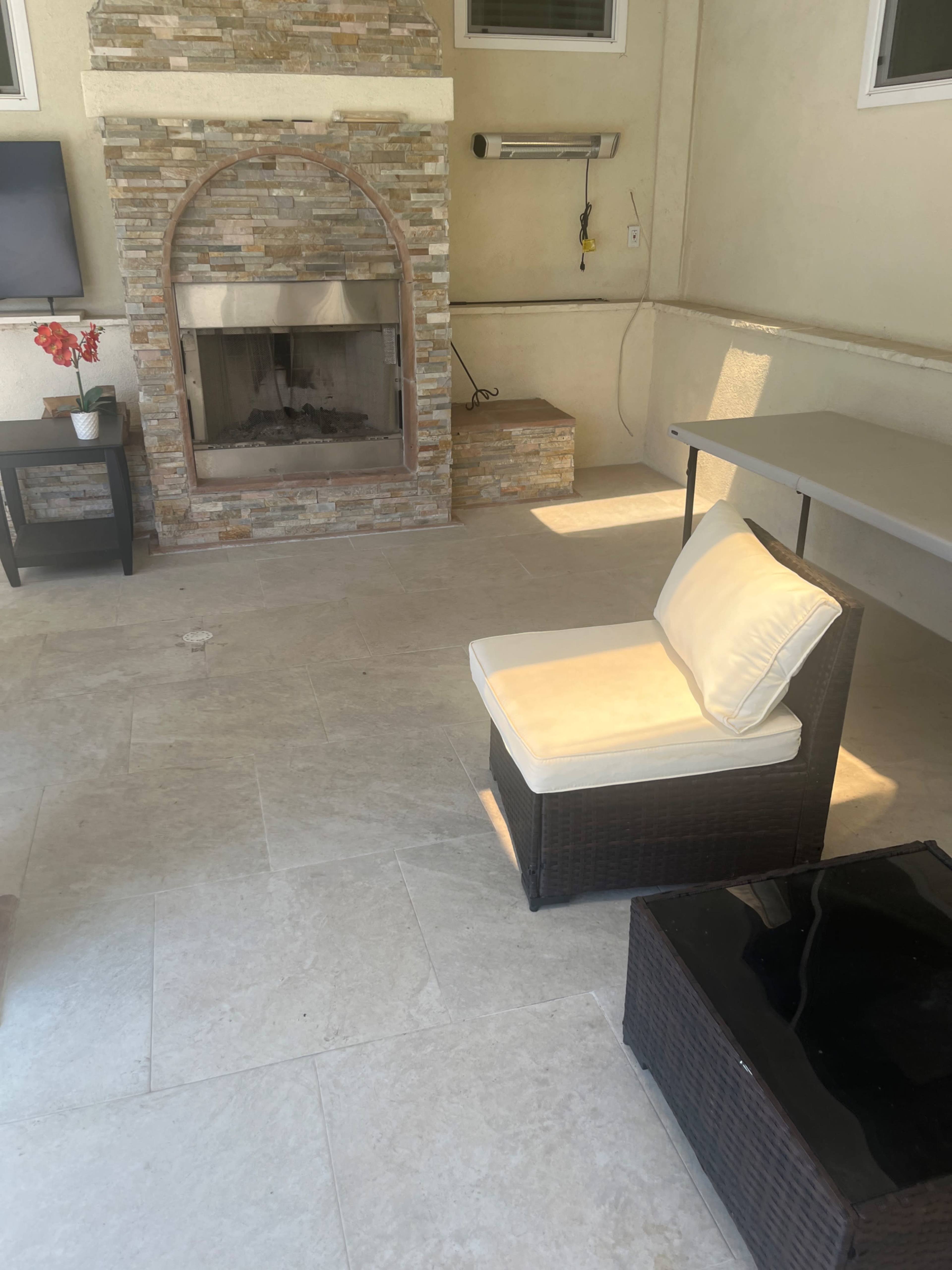 The image shows a covered outdoor seating area featuring a stone fireplace, a wicker chair with a cushion, and a small coffee table on a tiled floor.
