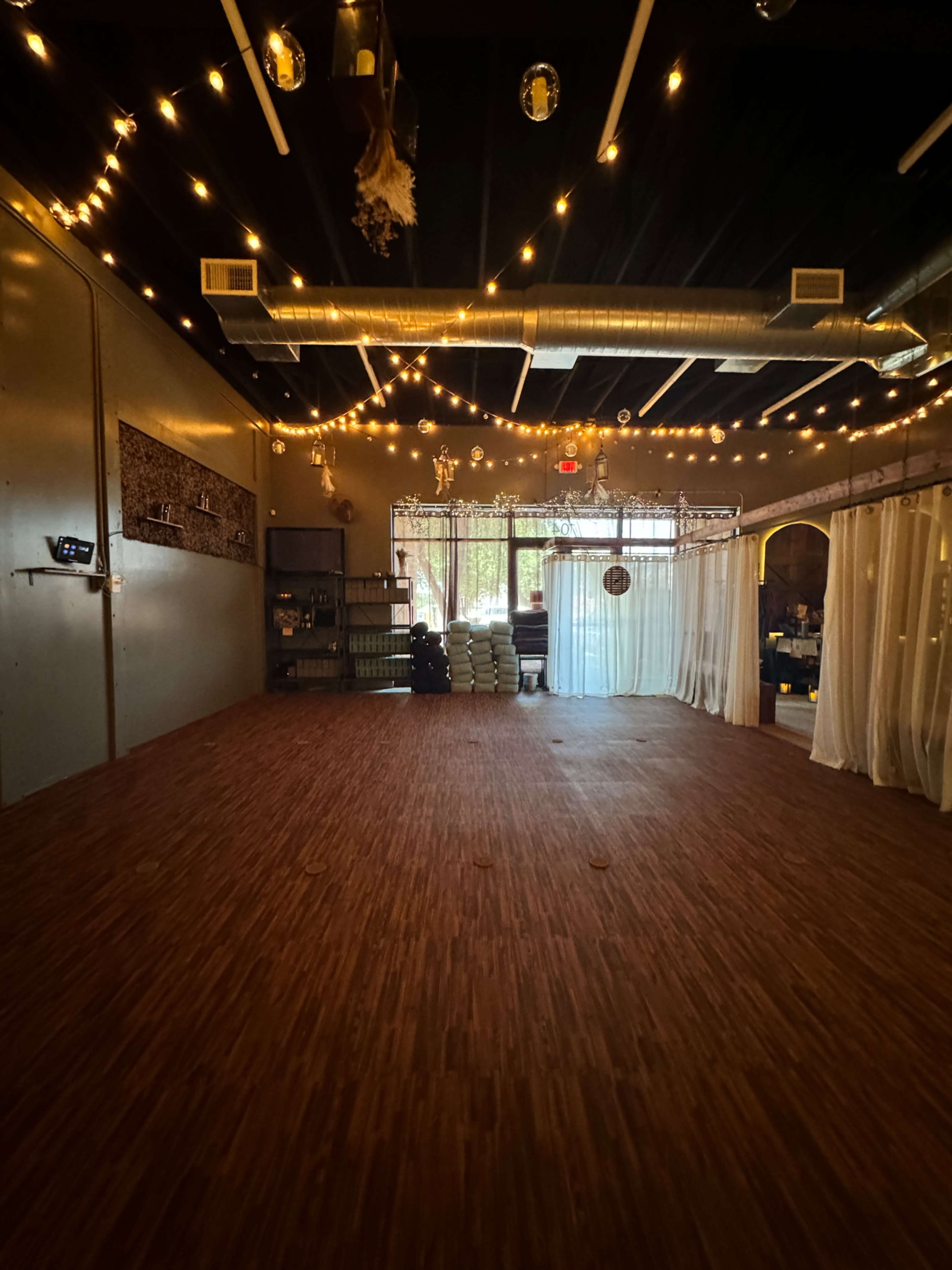 The image shows a spacious indoor studio with wooden flooring, draped curtains, and soft lighting from hanging bulbs.