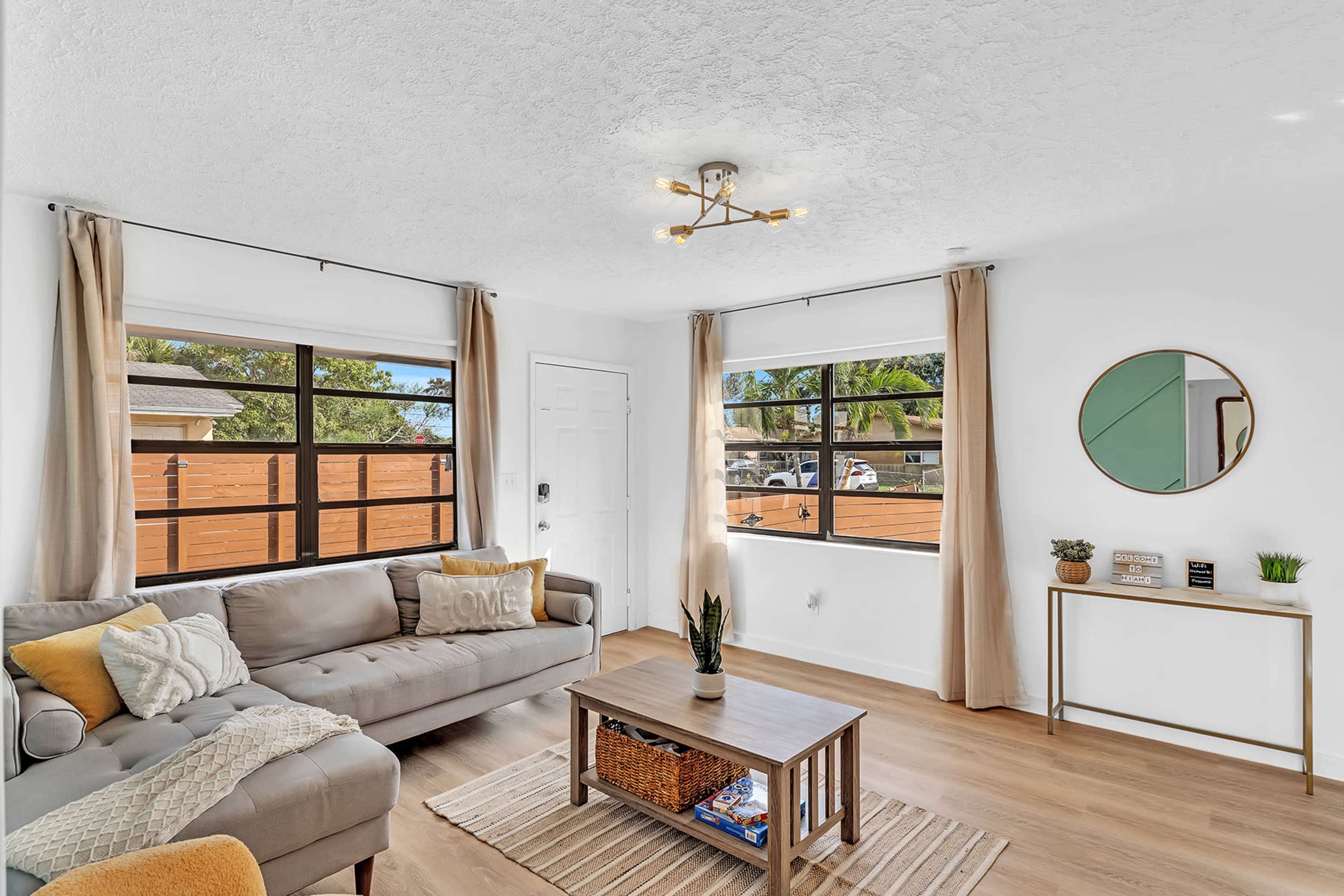 A bright living room featuring a gray couch, a wooden coffee table, and large windows with beige curtains.