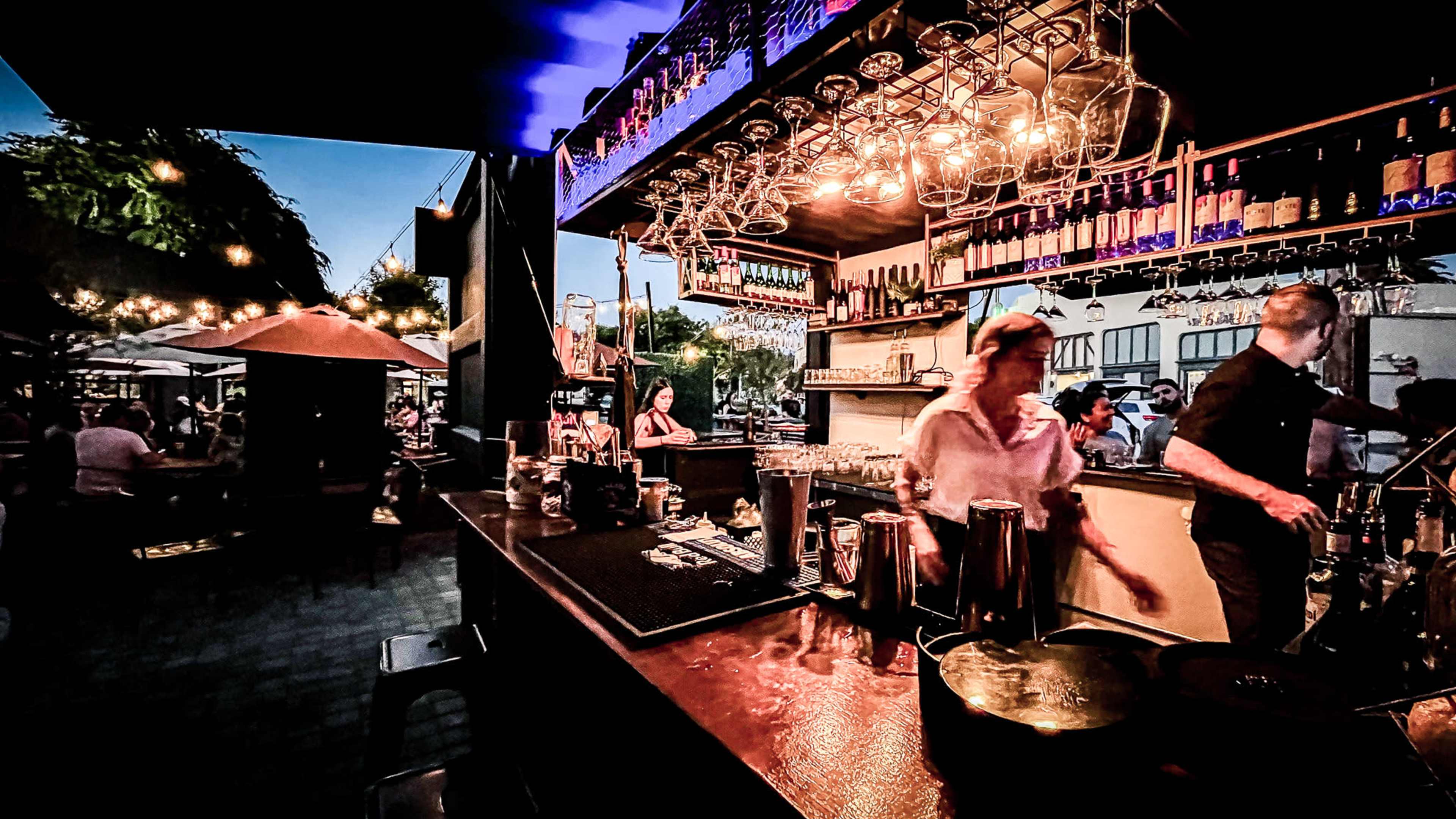 A bustling outdoor bar scene features a bartender preparing drinks while patrons sit at tables illuminated by string lights.