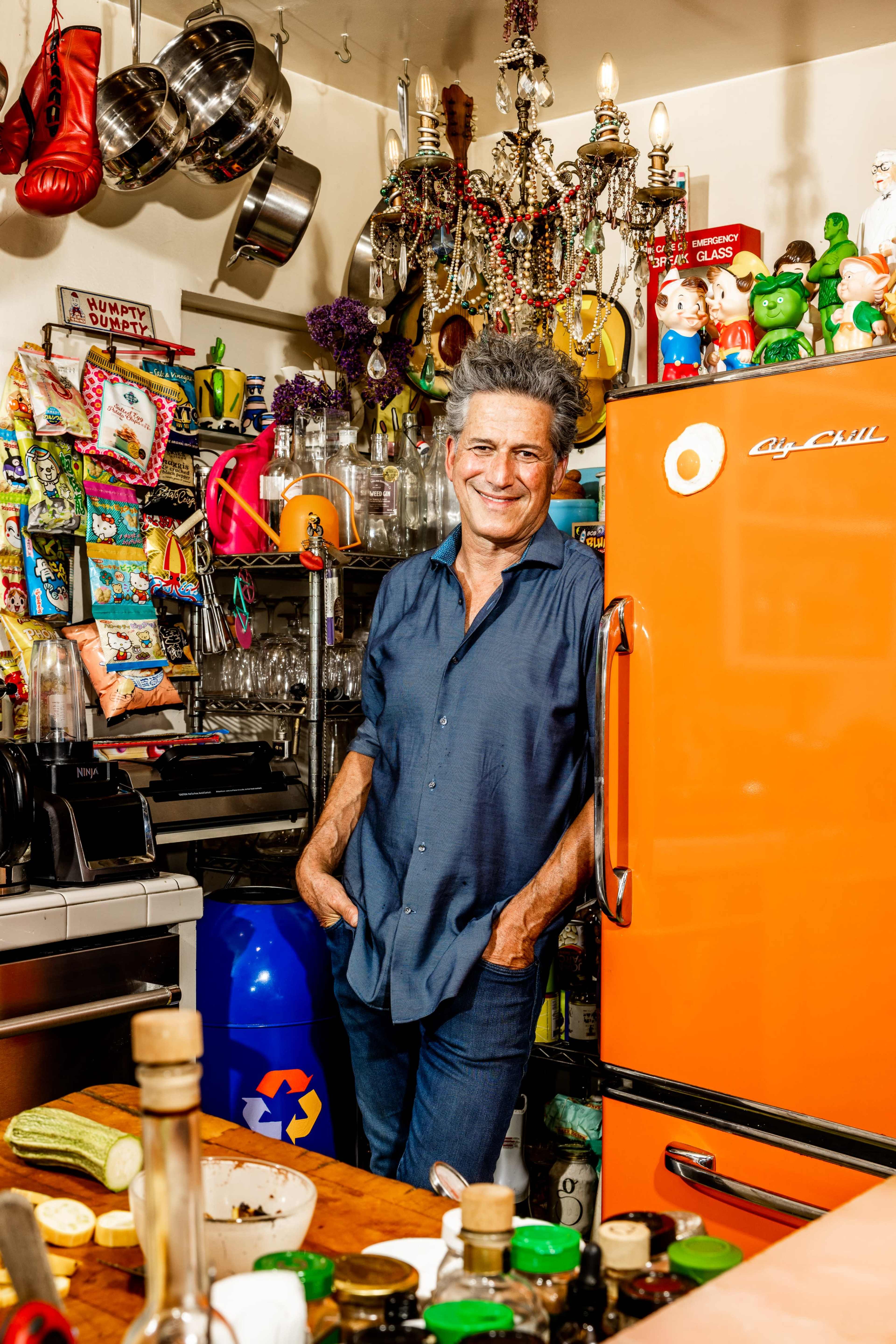 A man stands casually beside an orange refrigerator in a vibrant kitchen filled with colorful decor, utensils, and various kitchen items.