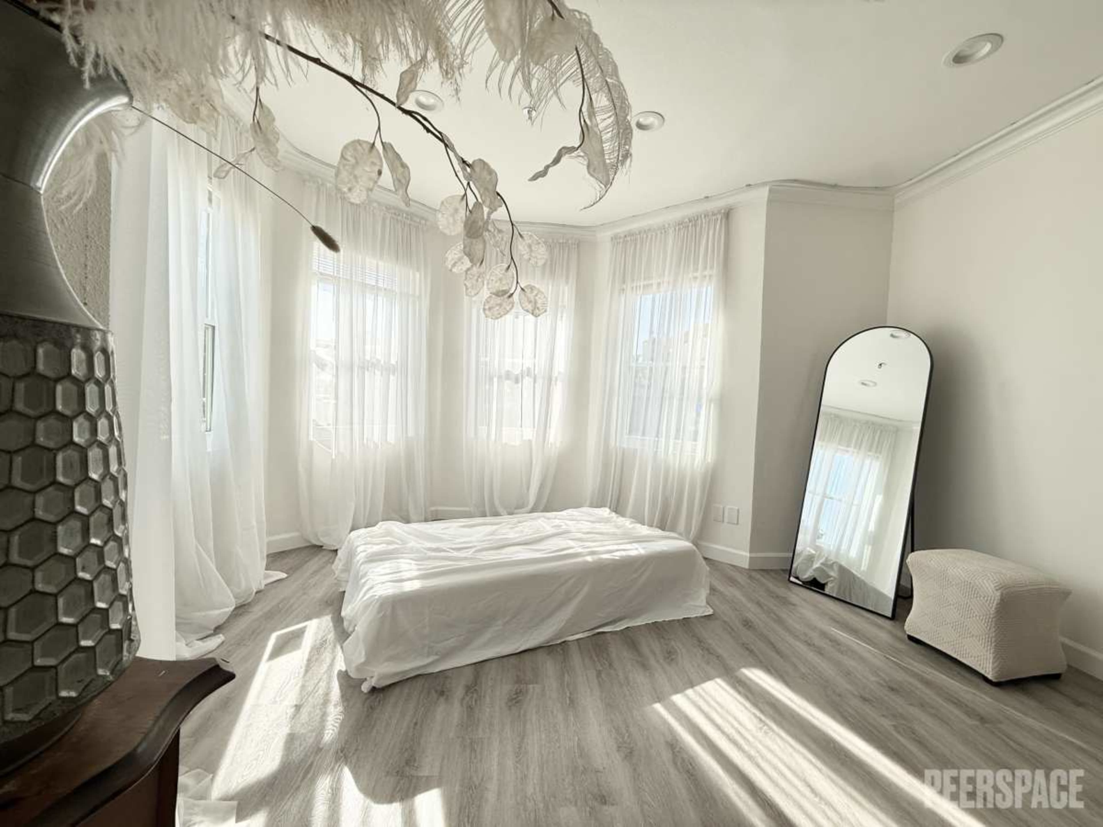 A bright room features large windows with sheer curtains, a minimalist bed with white bedding, and a floor-length mirror beside a small, upholstered chair.