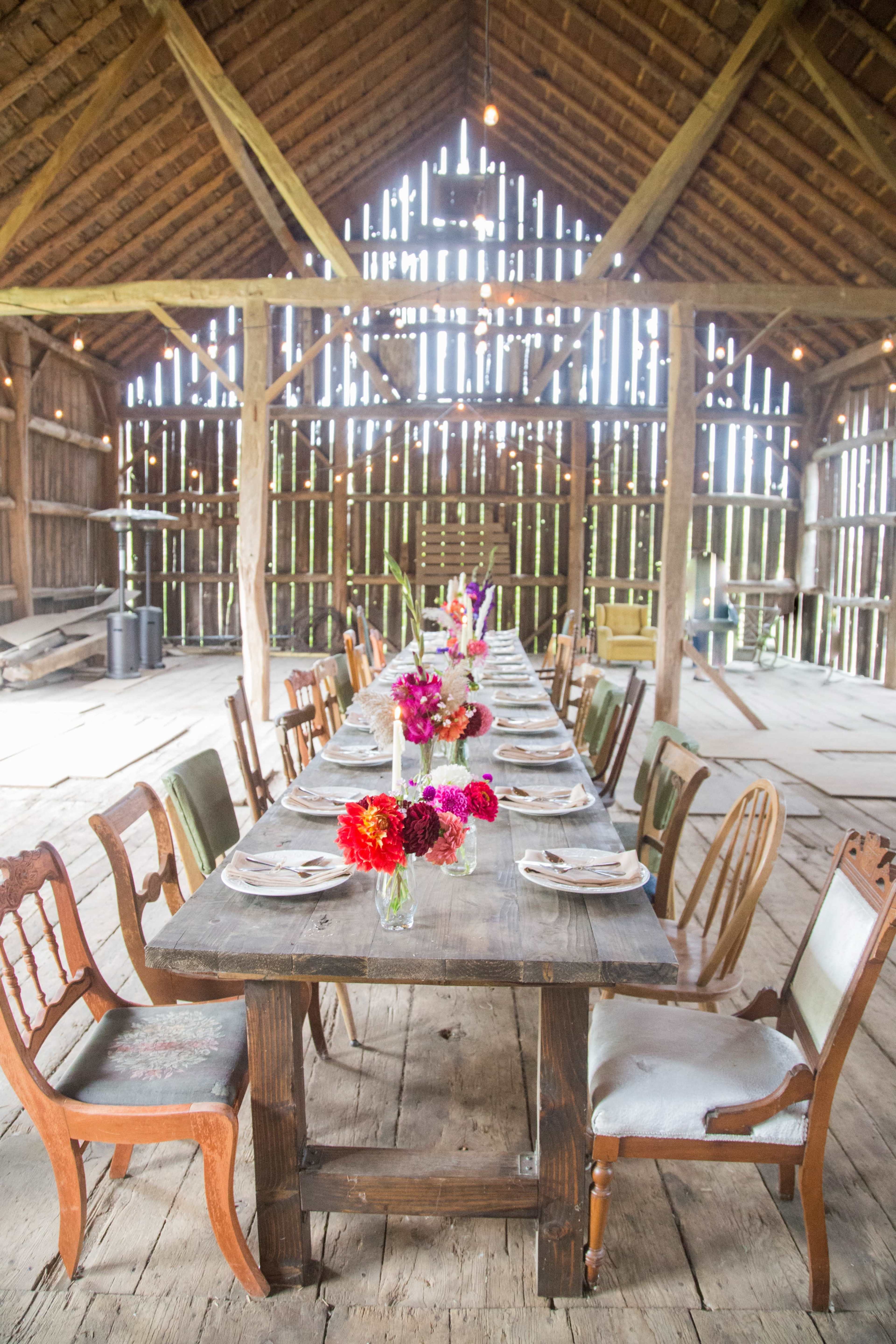 A long wooden table is set with plates and vibrant flower arrangements in a rustic barn with wooden beams and open slats.