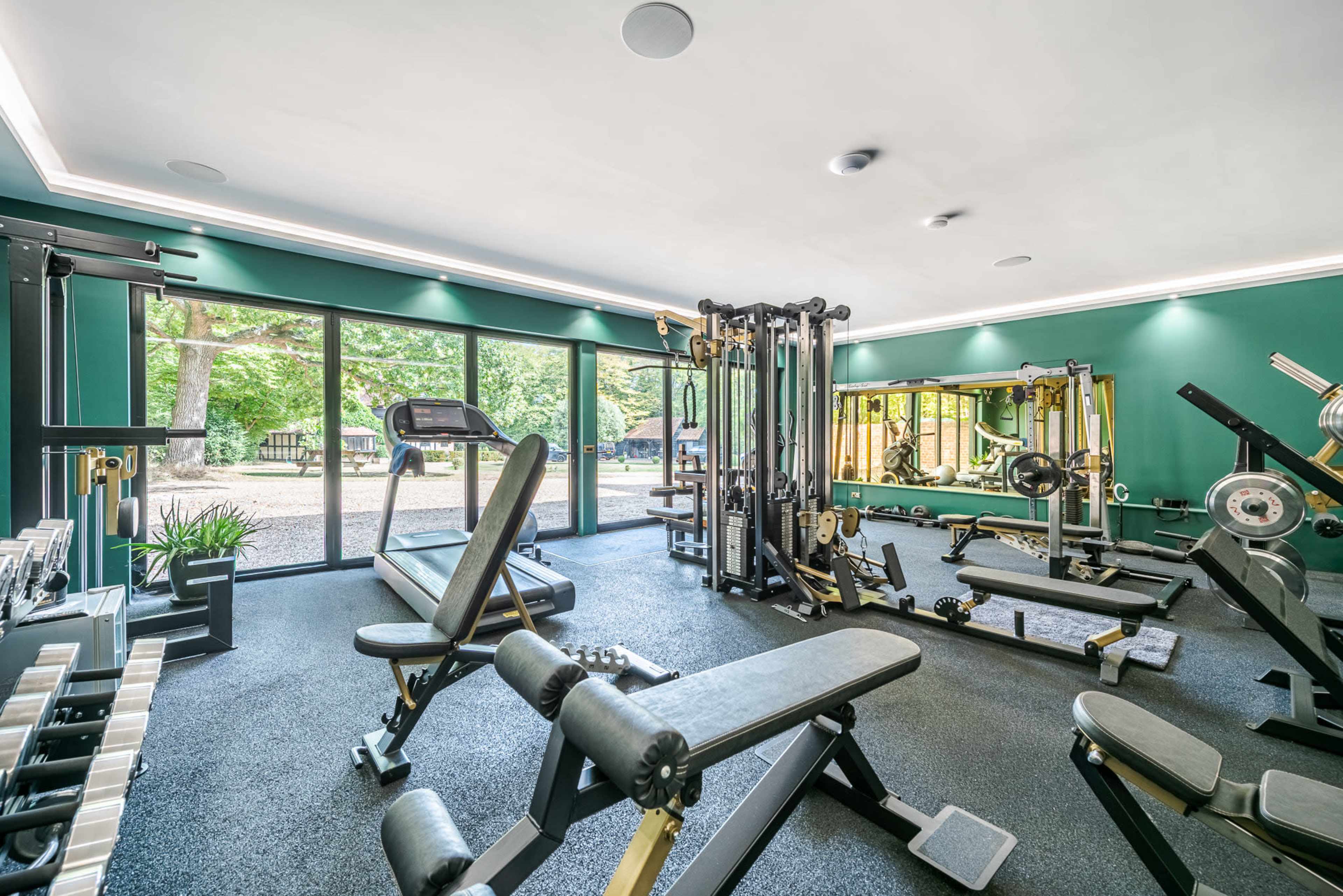 The image shows a well-equipped gym with various exercise machines, benches, and free weights, surrounded by large windows that offer a view of green trees outside.