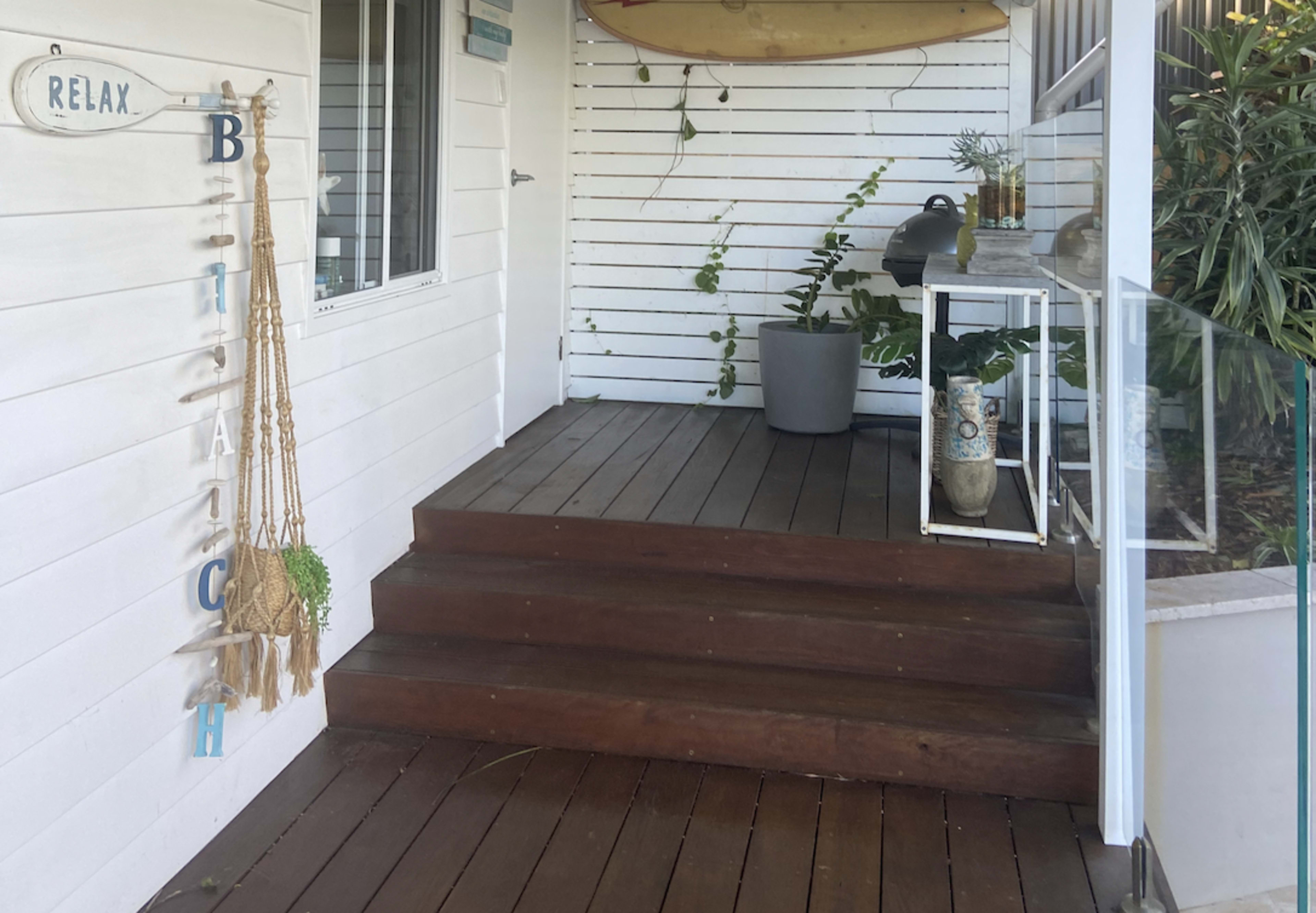 A wooden staircase leading to a white-paneled porch, with a surfboard leaning against the wall and plants in decorative pots.