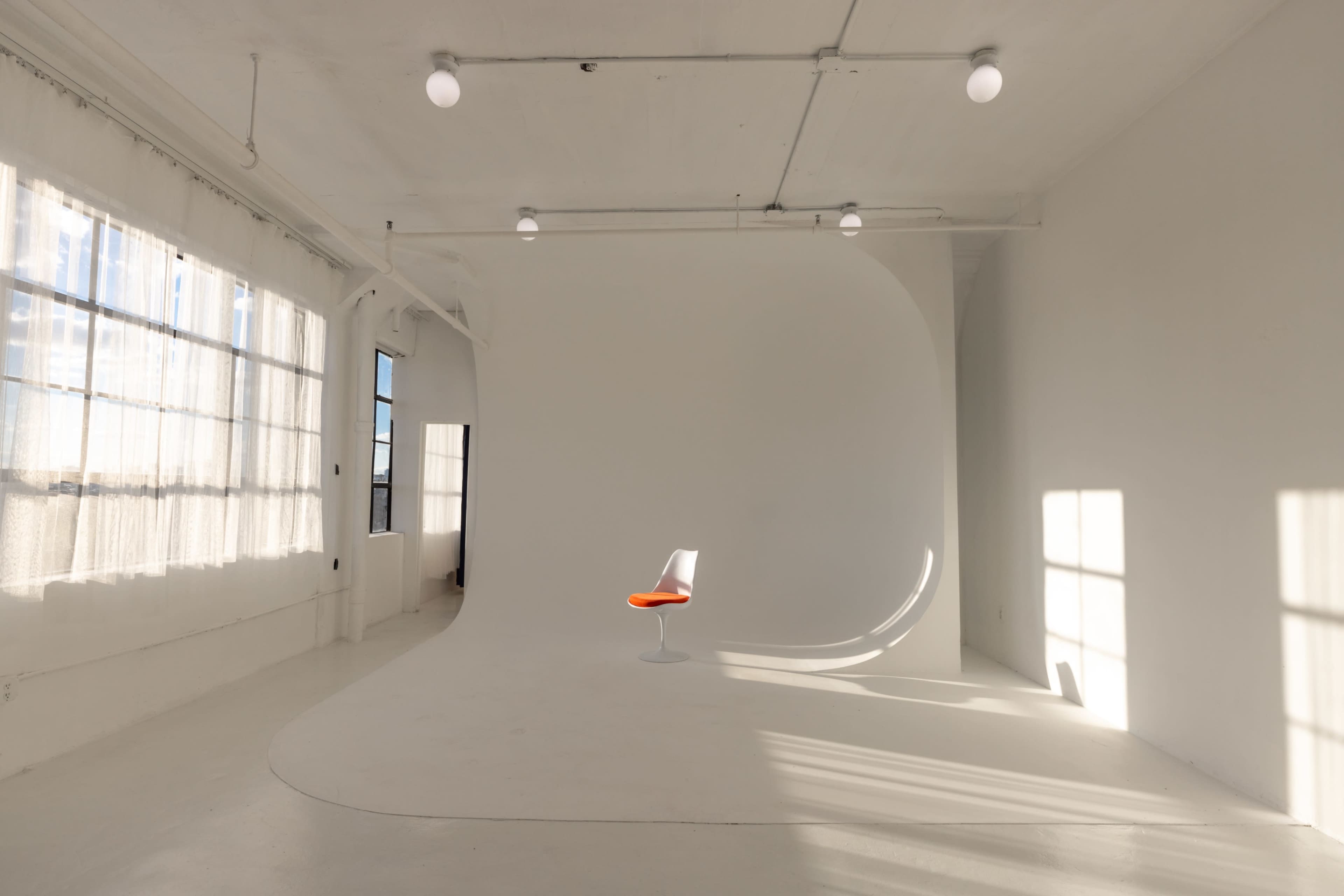 A bright, minimalistic studio space featuring a single white chair with an orange seat placed against a curved white backdrop.