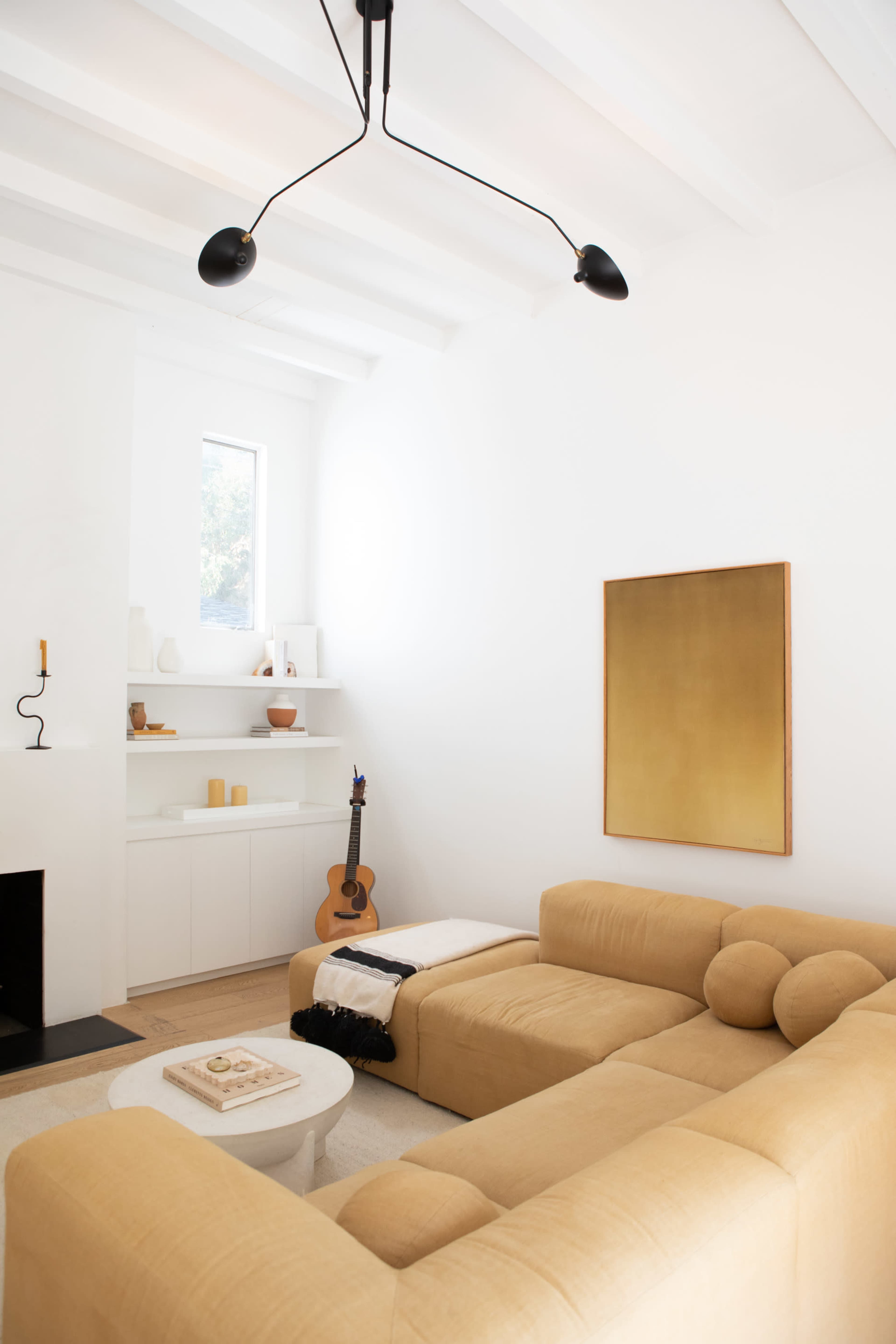 A spacious living room features a beige sectional sofa, a white coffee table, a wooden guitar, and a gold-framed painting on the wall.