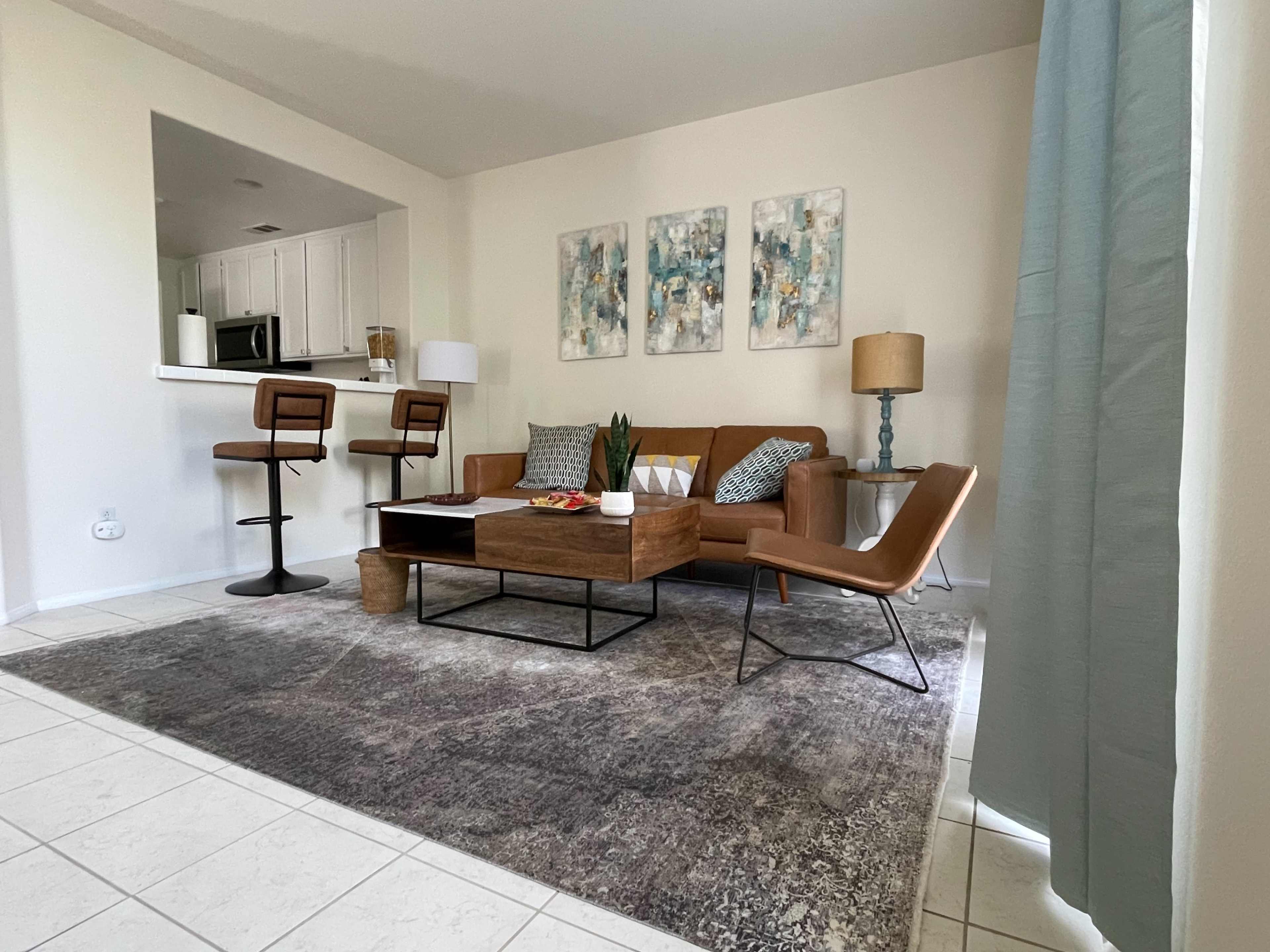A living room with a tan leather sofa, a wooden coffee table, two bar stools at a kitchen counter in the background, and abstract wall art.