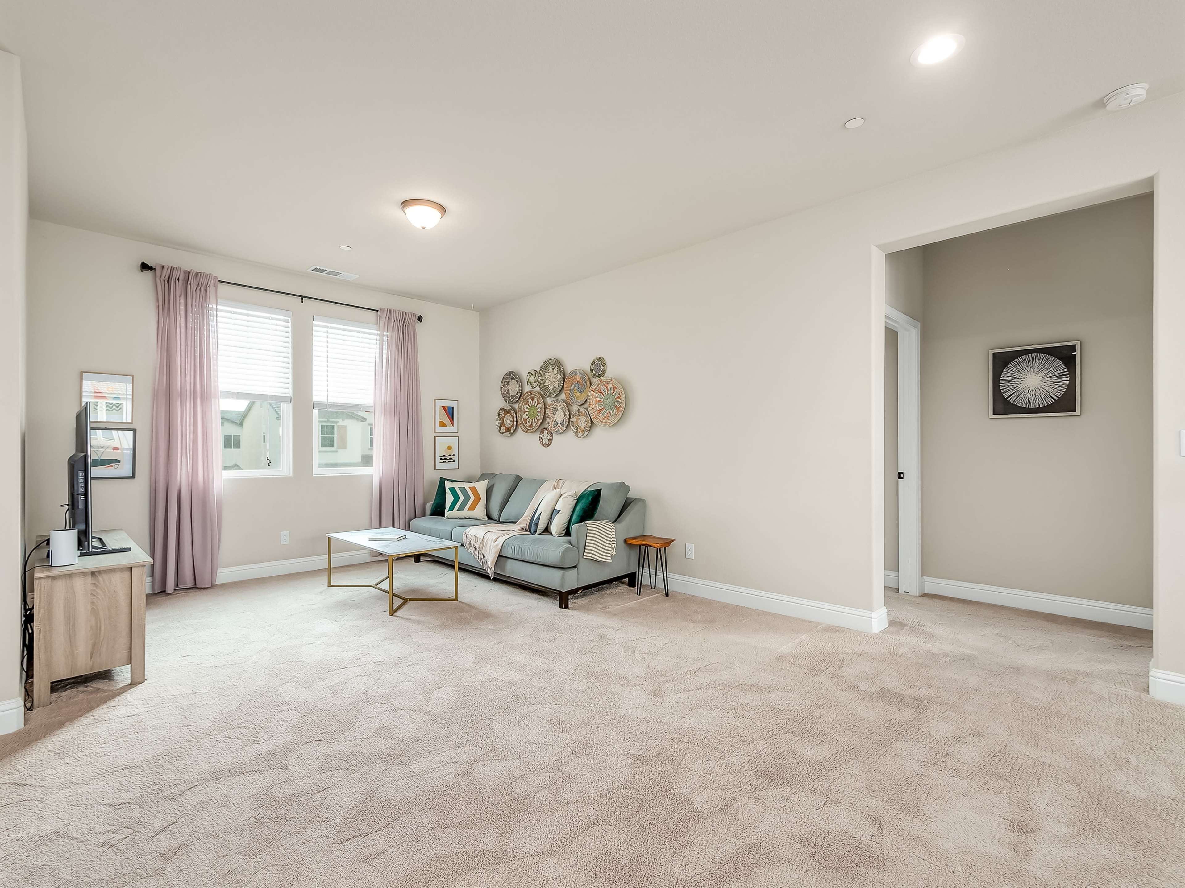 A spacious living room with a light carpet, a sofa adorned with colorful cushions, a coffee table, and wall decor, complemented by natural light from the two windows.