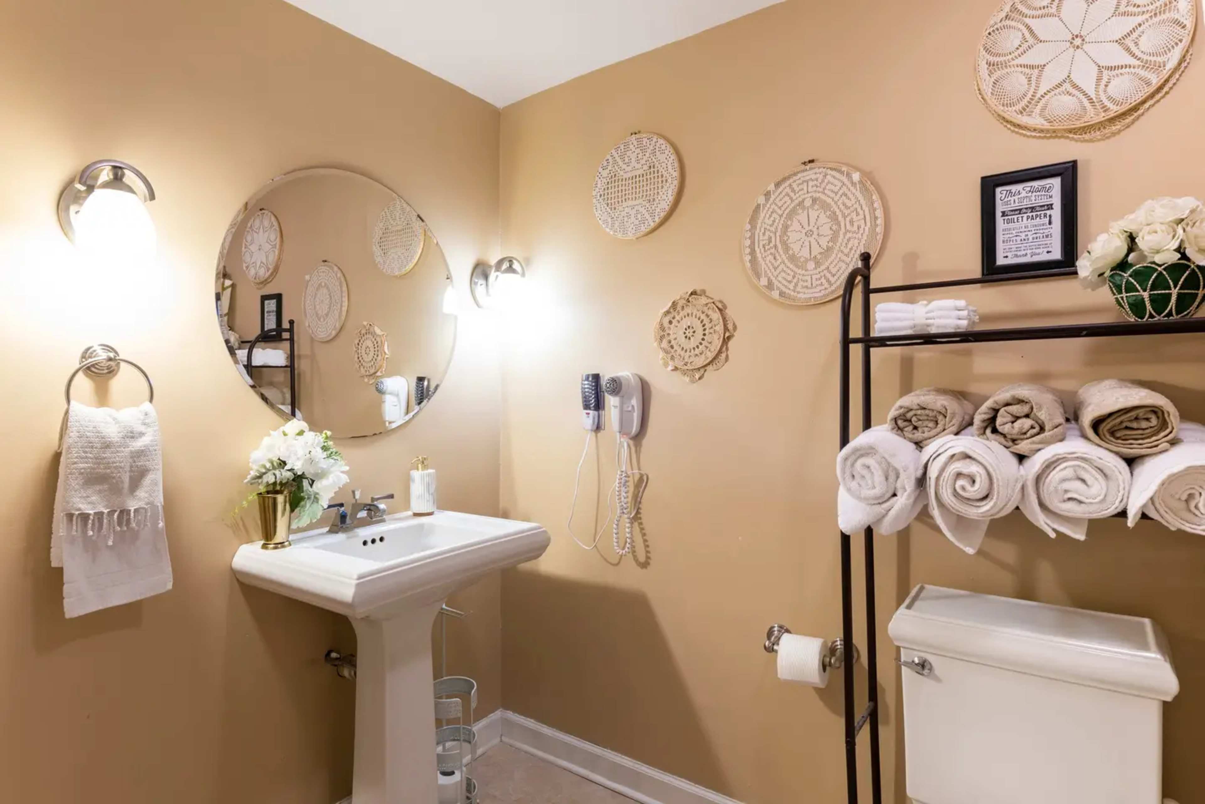 The image shows a bathroom with a round mirror above a white sink, beige walls adorned with decorative wall hangings, and neatly arranged folded towels on a rack.
