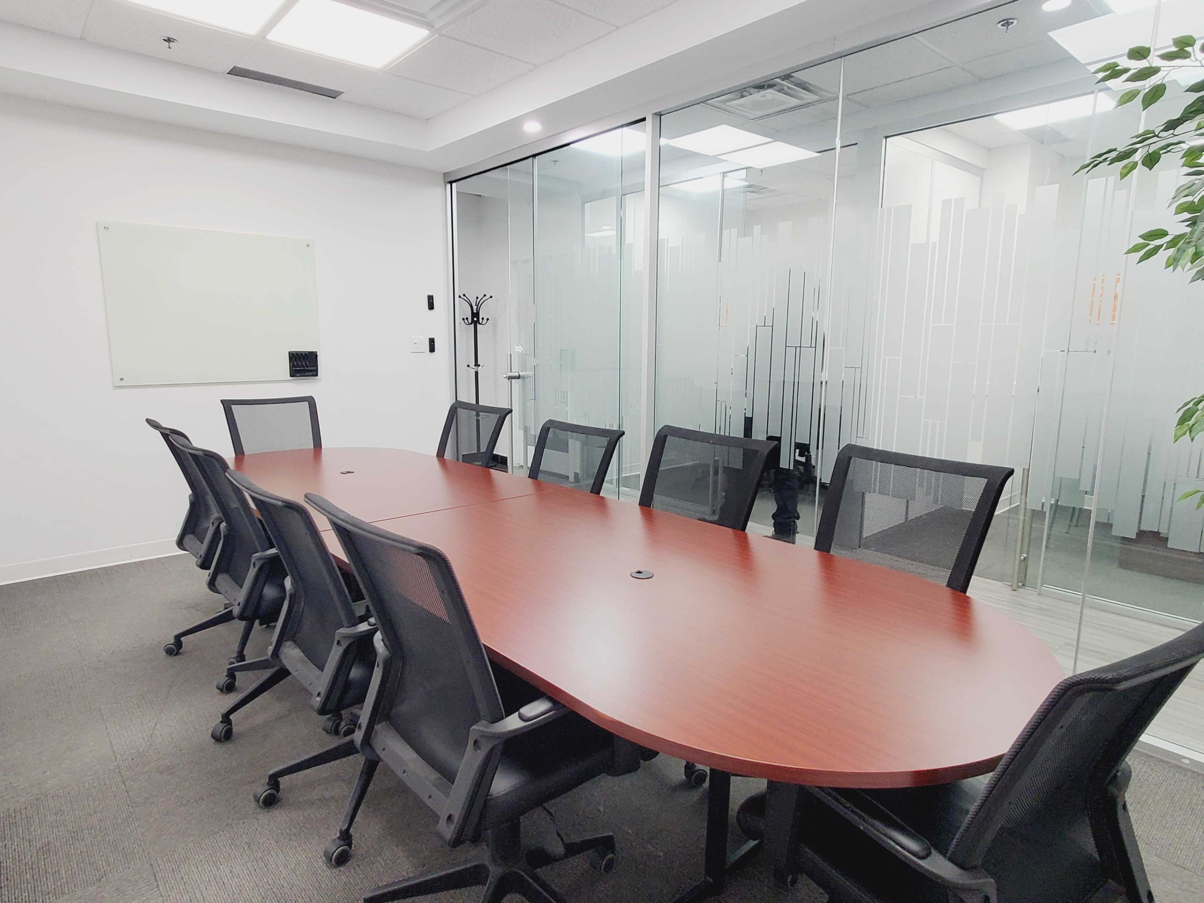 A conference room features a long oval wooden table surrounded by ergonomic black chairs and a glass wall with a frosted design.