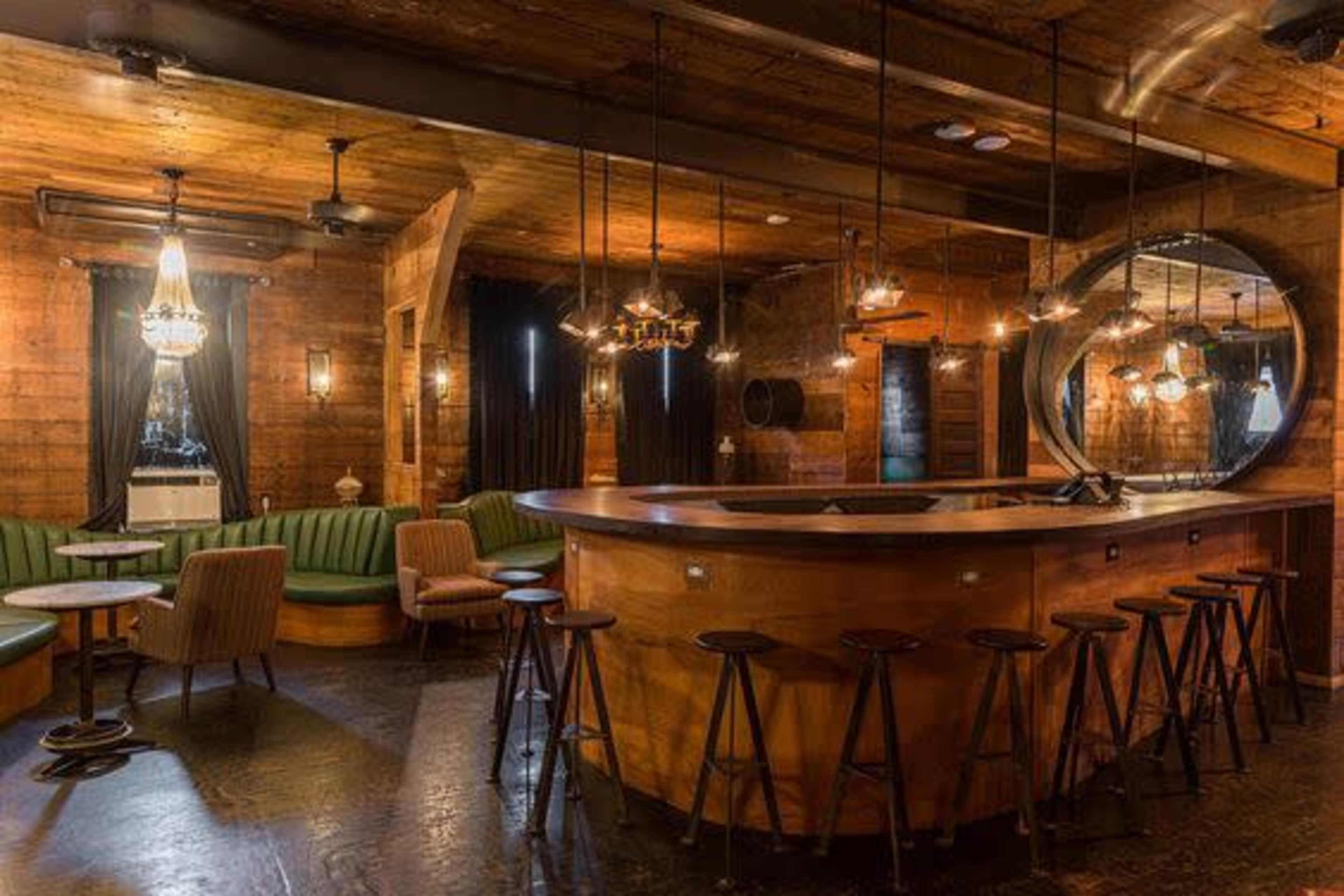 A wood-paneled bar interior with a curved wooden counter, green upholstered seating, and hanging light fixtures creating a cozy atmosphere.