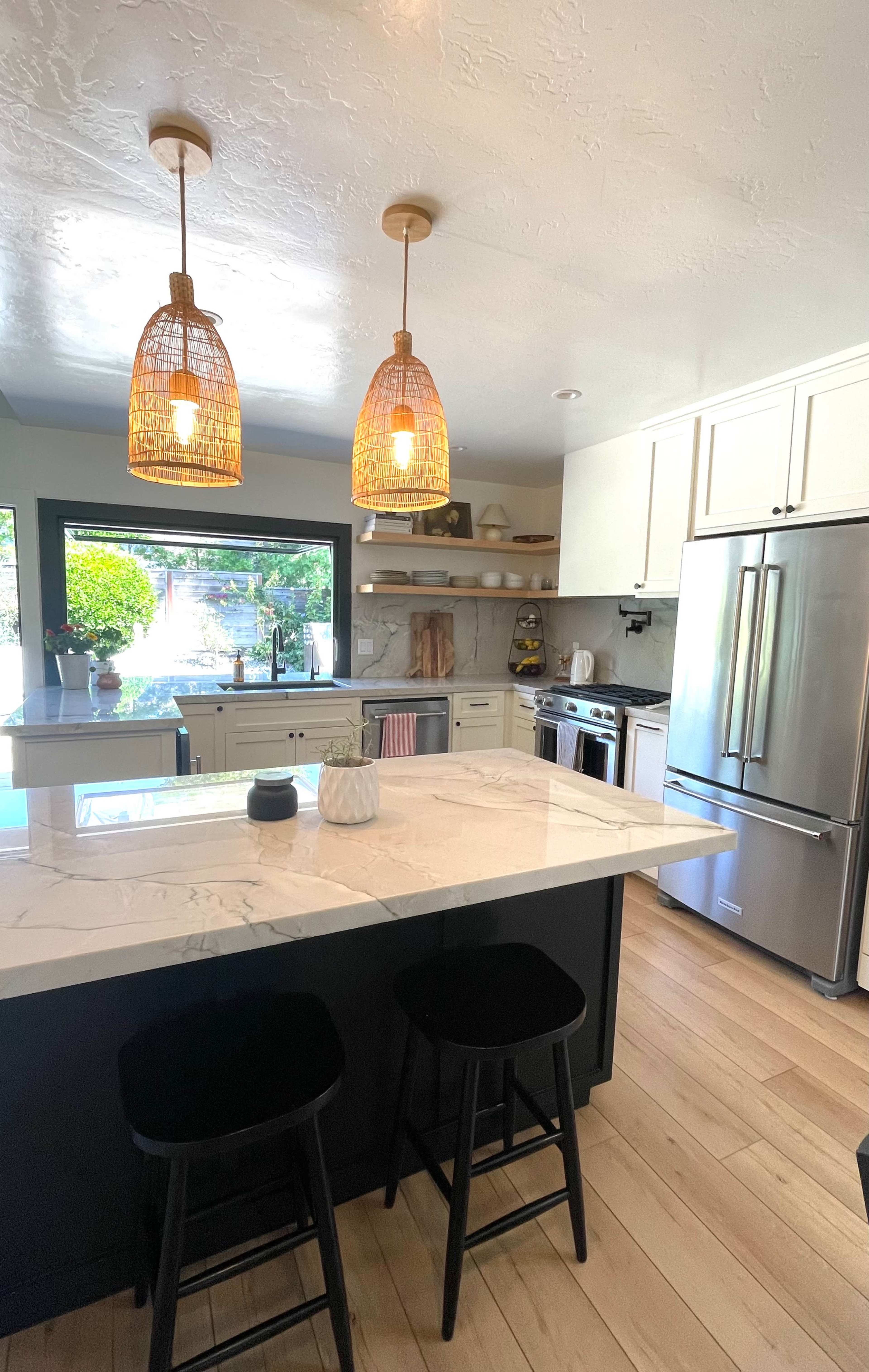 A modern kitchen features a large island with two black stools, pendant lights overhead, and stainless steel appliances.