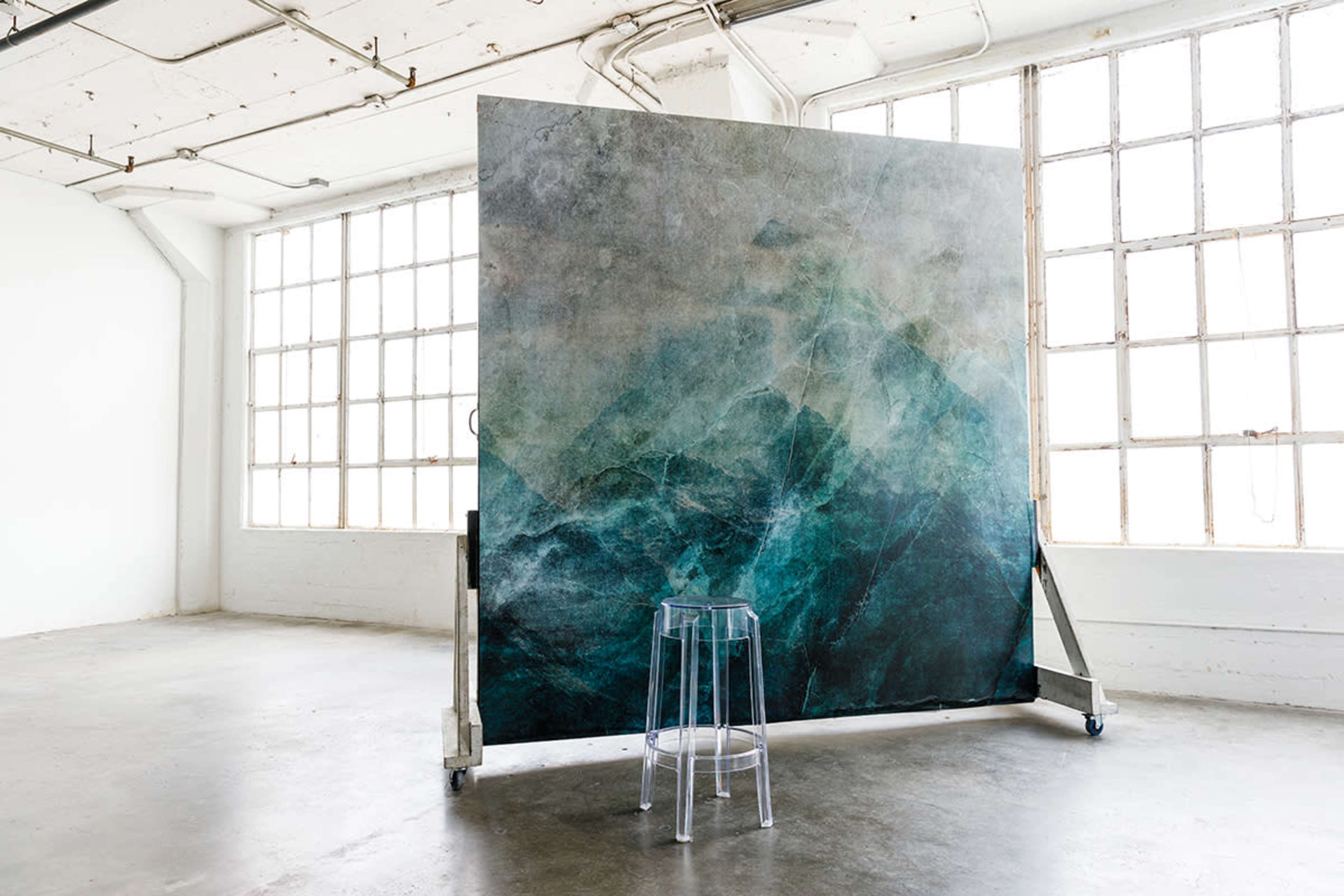 The image features a large abstract backdrop with teal and gray tones, positioned in a bright, minimalist studio space with large windows.