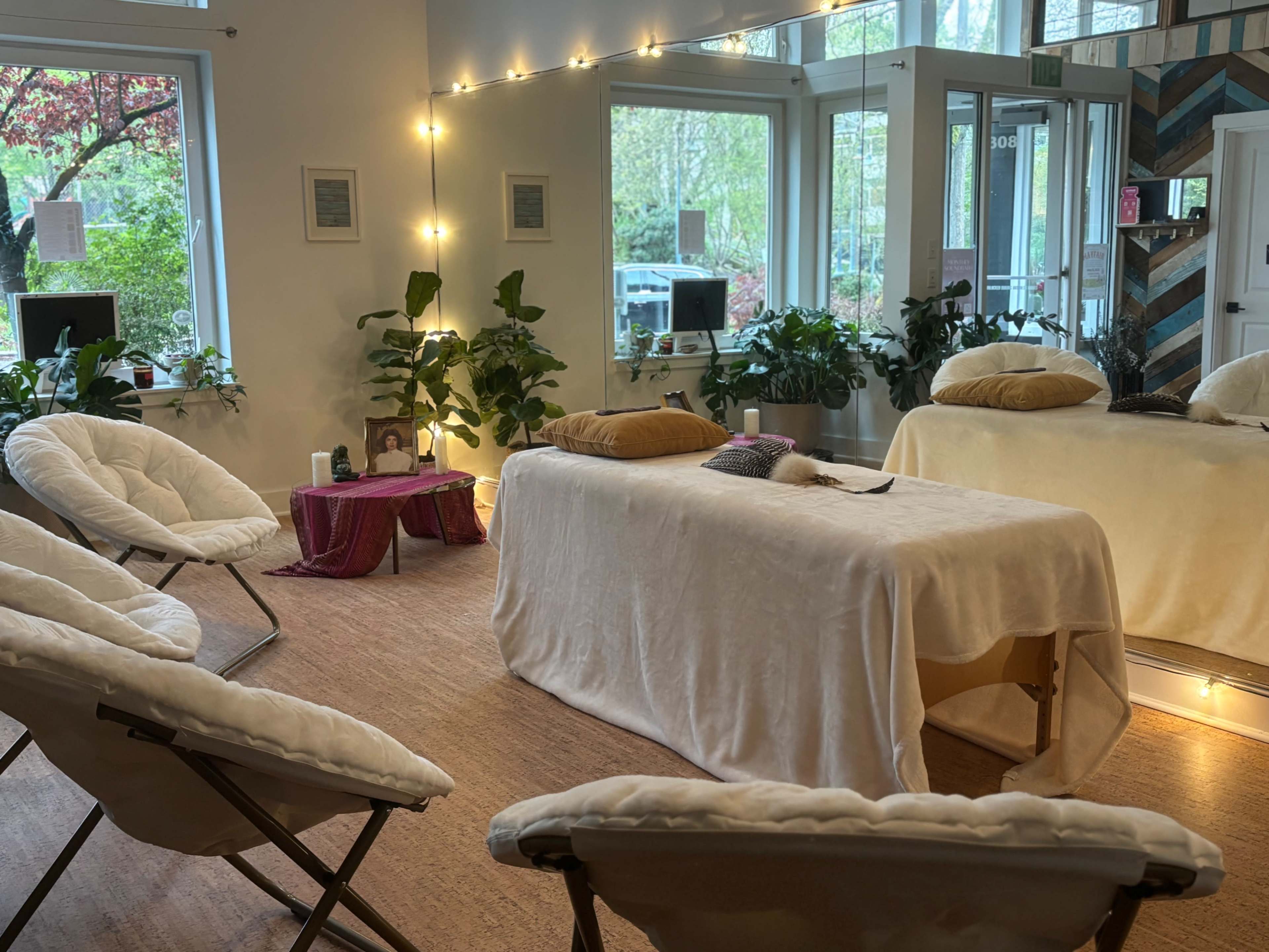 The image shows a serene room with several folding chairs arranged around a massage table, surrounded by indoor plants and soft lighting.