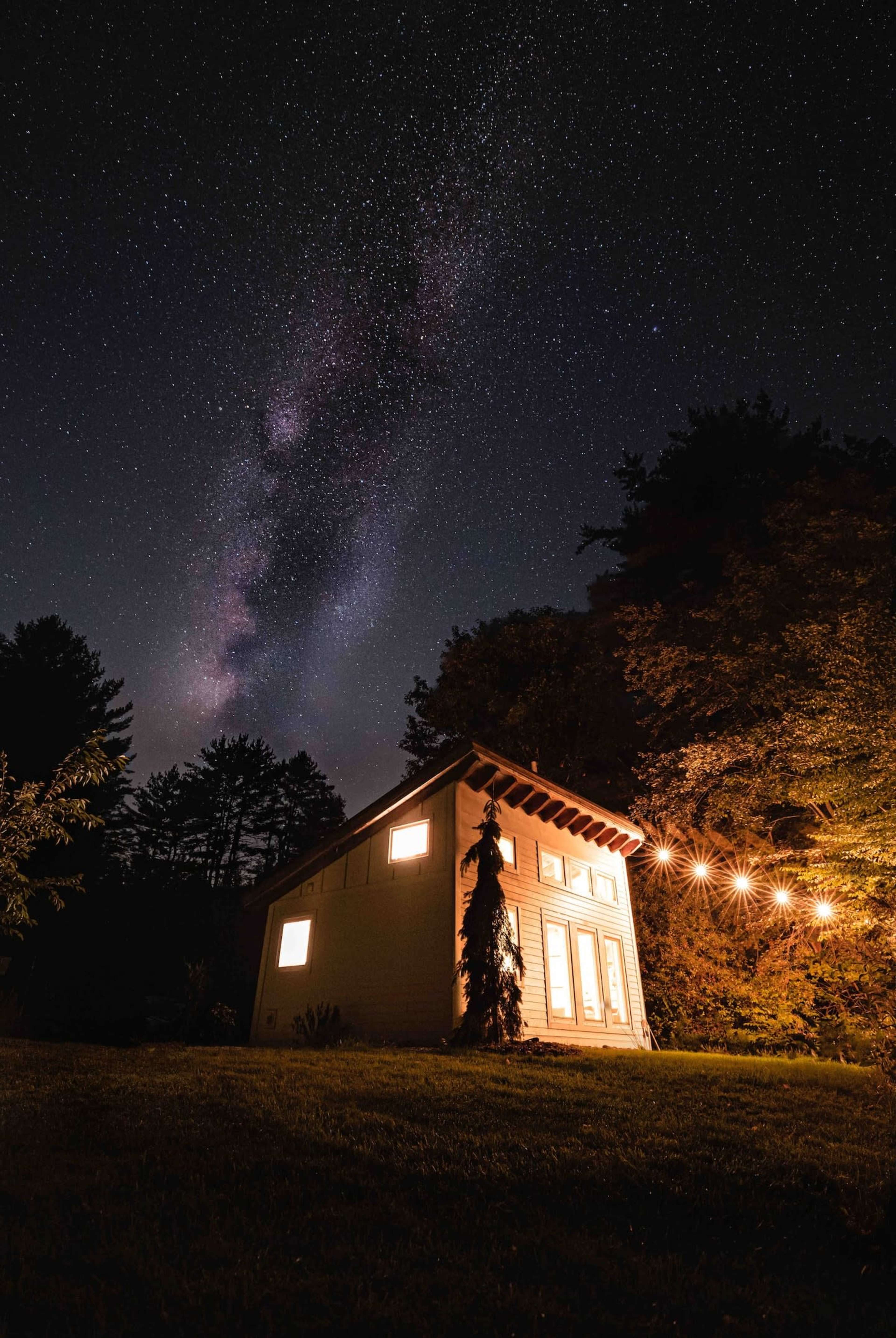 A small house is illuminated by warm lights under a starry night sky with the Milky Way visible above.