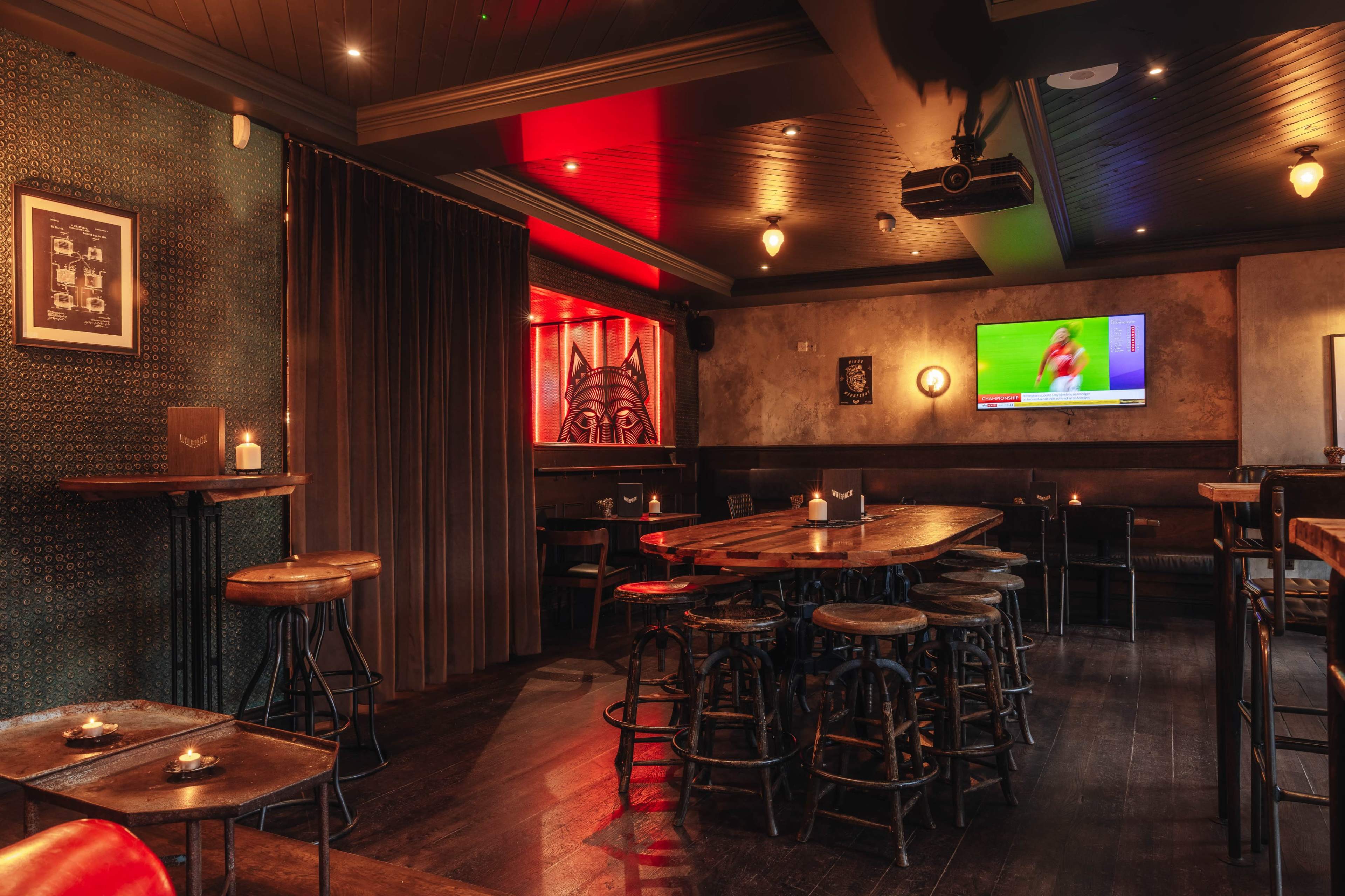 The image shows a dimly lit indoor bar with wooden tables and stools, a large screen displaying a sports game, and red and green accent lighting.